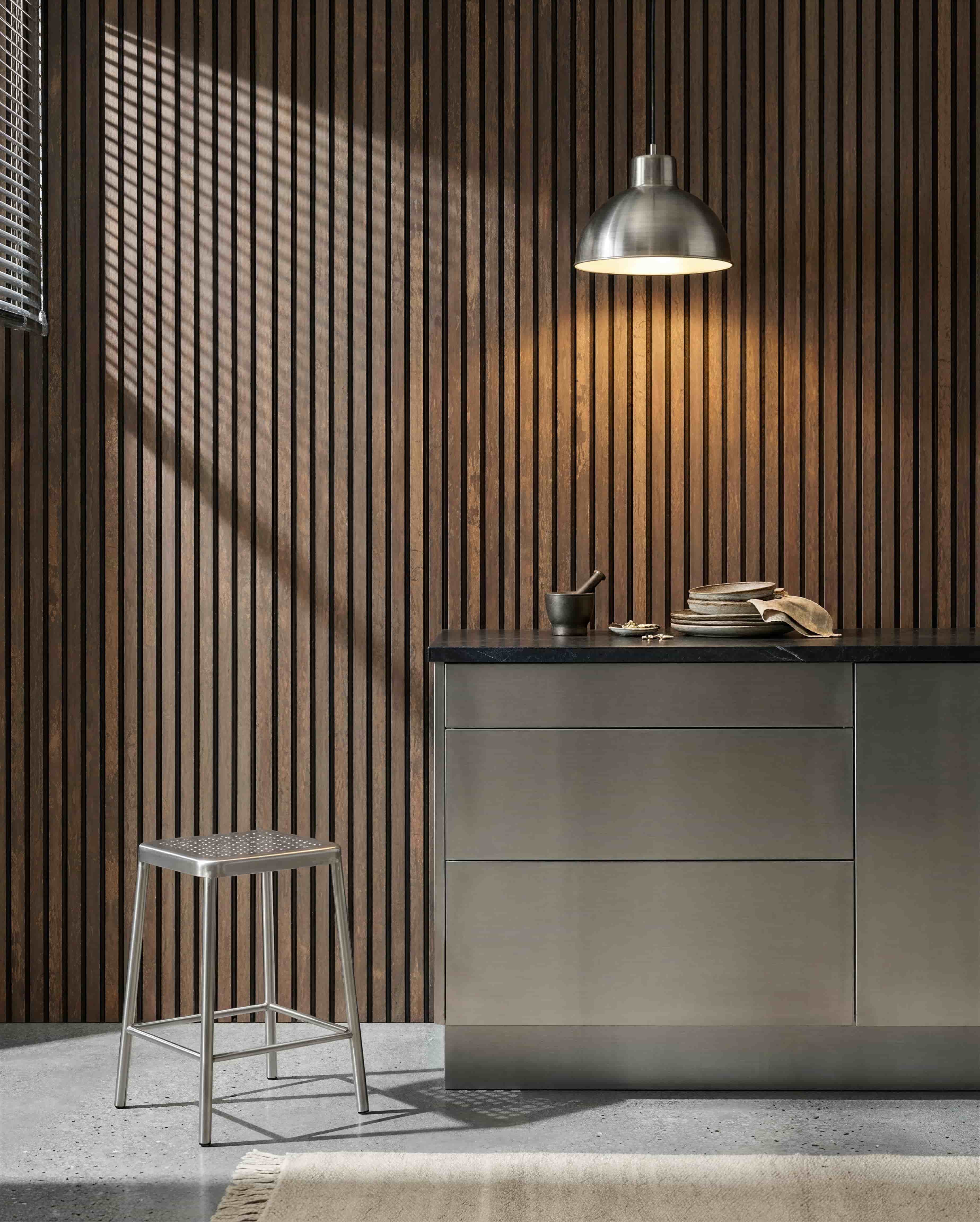 Küche mit einer WoodUpp Akupanel Oxide Copper Akustik-Lamellenwand, Edelstahl-Arbeitsplatte, Metallhocker, Pendelleuchte, verschiedenen Geschirrteilen sowie Mörser und Stößel.