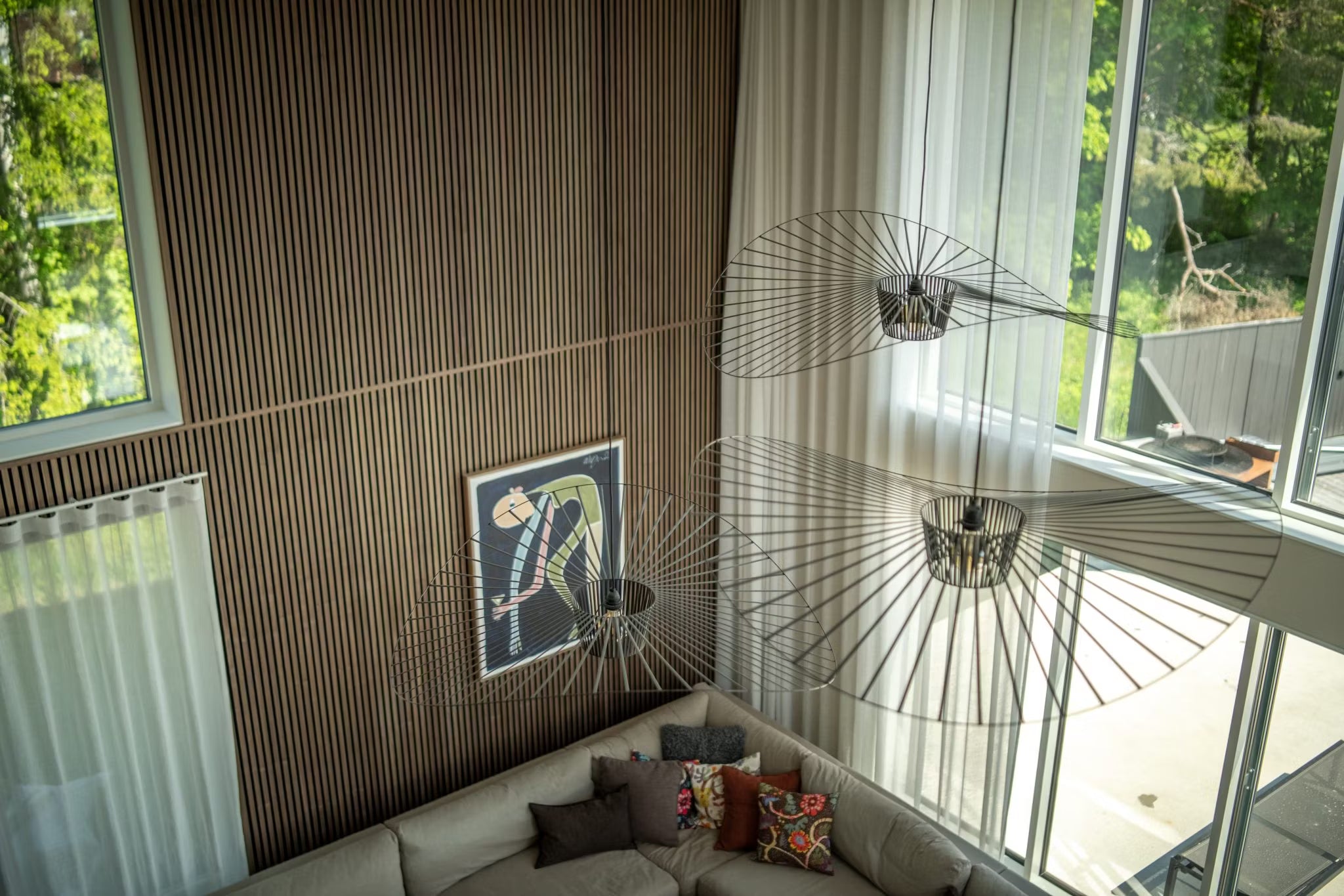 Modernes Wohnzimmer mit Akupanel-Wänden aus nordischem Holz, großen Fenstern, abstrakter Akustikpaneel-Kunst, einem Sektionssofa und minimalistischen Drahtgitter-Leuchten, von oben gesehen.