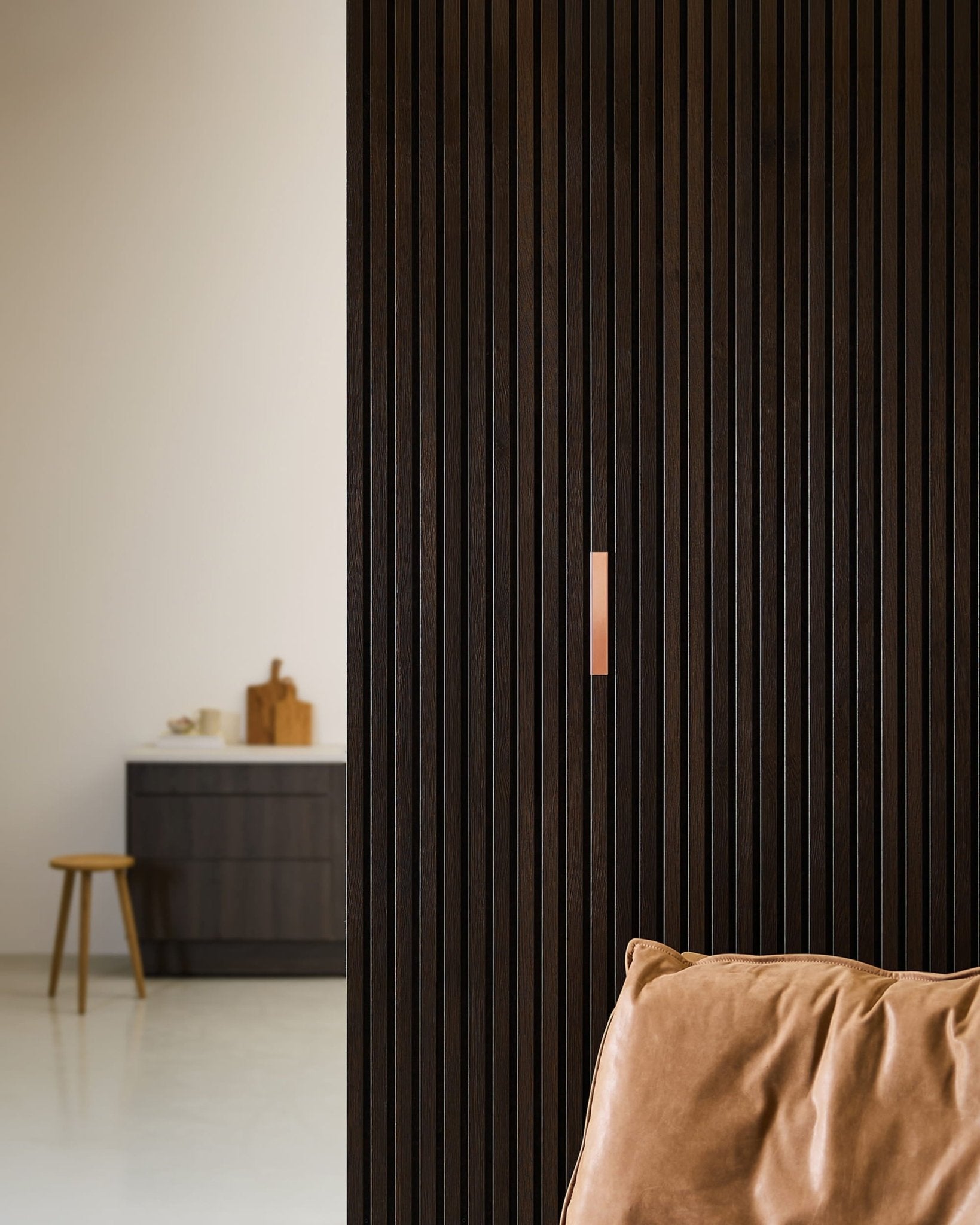 Minimalistisches Interieur mit einem braunen Ledersessel, WoodUpp Liva | Cover / 20 cm breiten Akustikpaneelen in Kupferoptik an der Wand sowie einem Hocker an einer Theke mit Schneidebrettern.