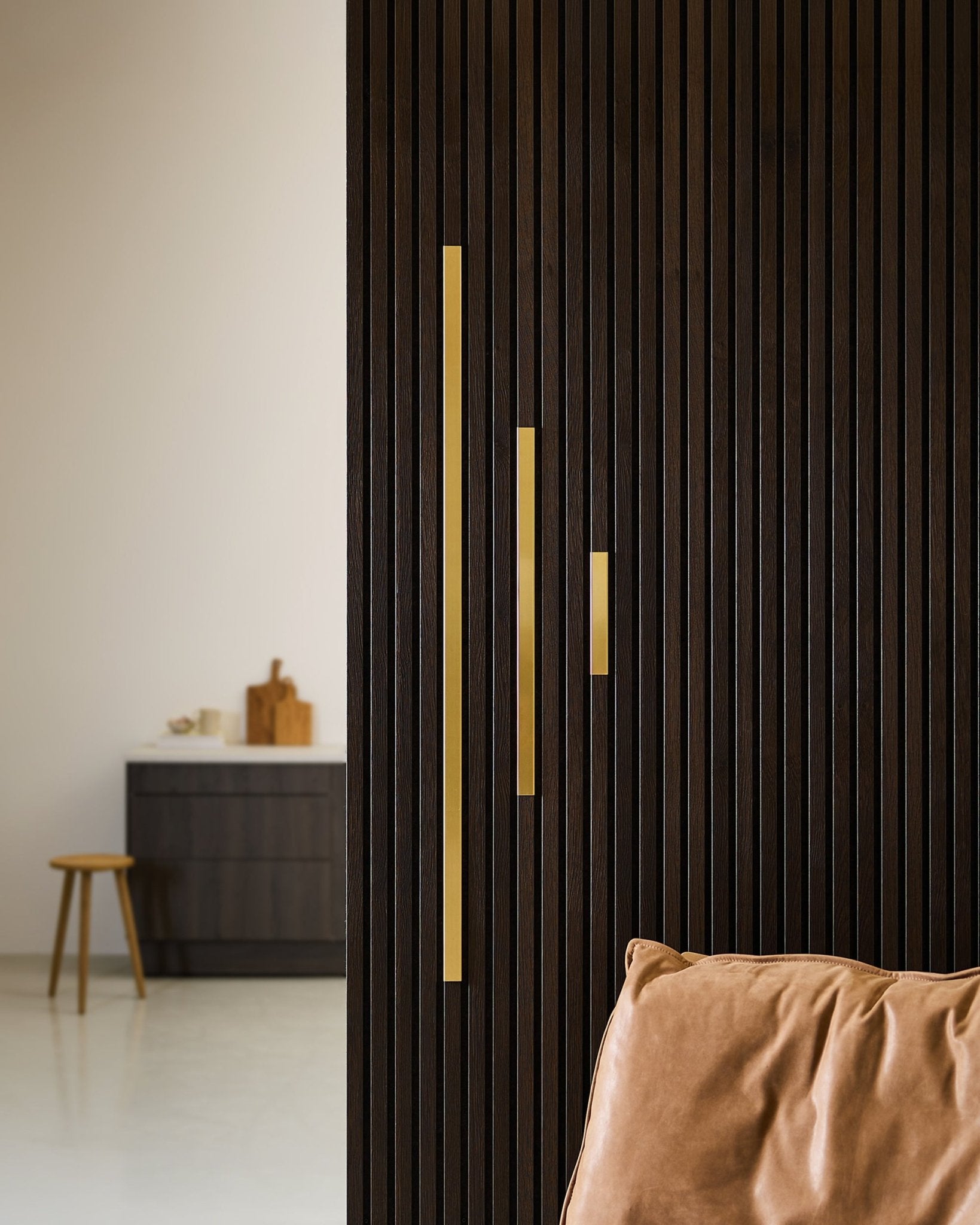 Modernes Interieur mit einer mit brass akzentuierten Akustik-Lamellenwand von WoodUpp, goldenen Griffen, einem sandfarbenen Kissen, einem Holzstuhl und einem dunklen Schrank.