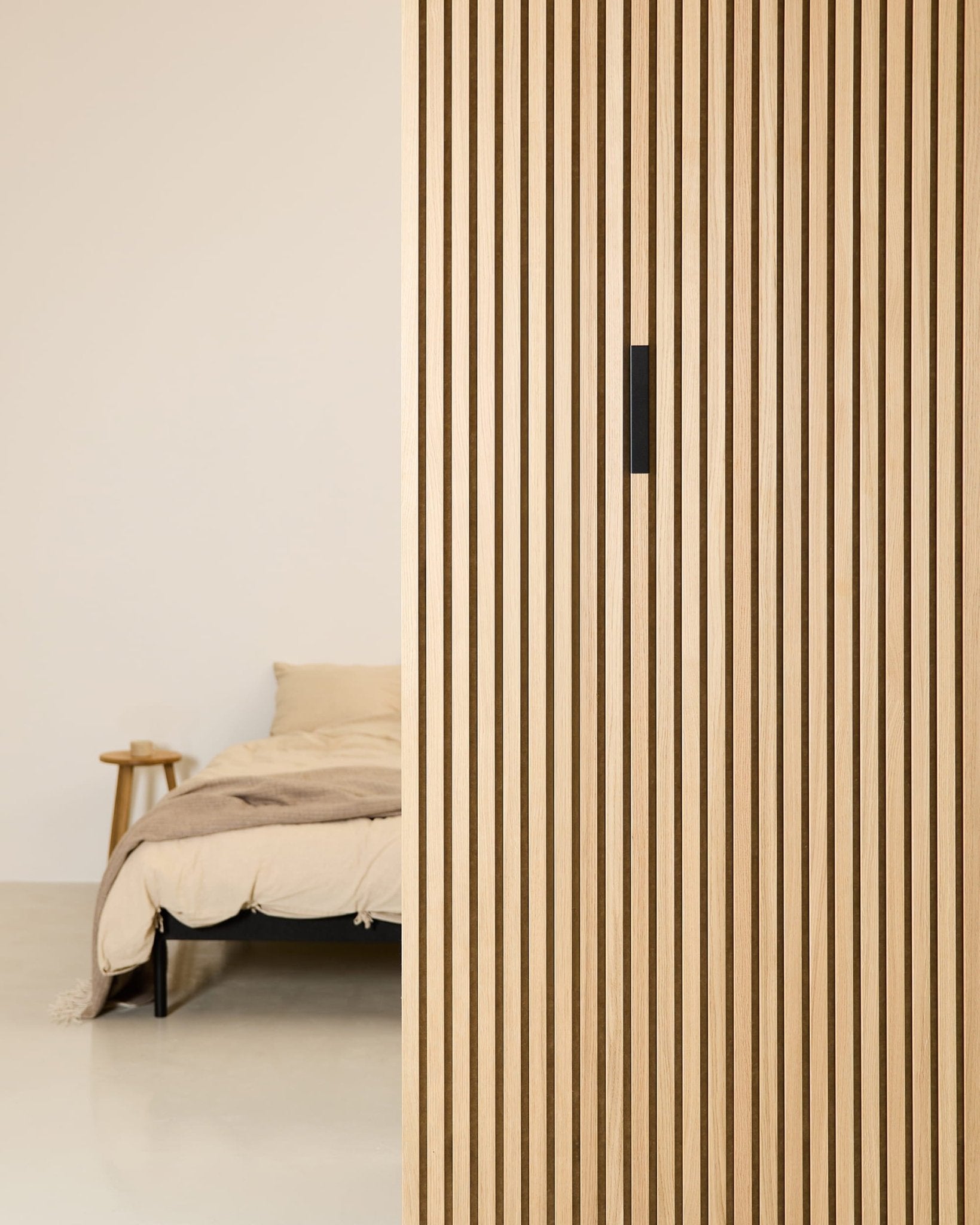 Minimalistisches Schlafzimmer mit einem beigen Bett, einem Beistelltisch aus Holz und einer Akustikplatte WoodUpp Liva Black Aluminum, die der vertikalen Trennwand Struktur verleiht.