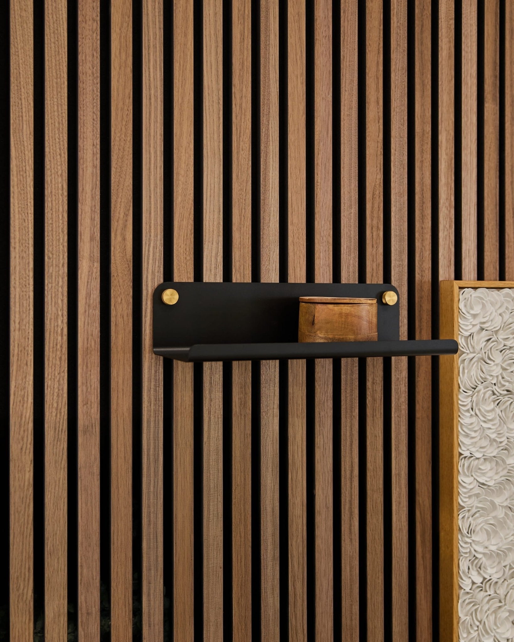 Eine kleine Holzkiste auf einem WoodUpp Norr Wide Regal (schwarz, 35,9 cm), das an vertikalen Akupanel-Akustikholzlamellen befestigt ist, wobei eine strukturierte weiße Platte zu sehen ist.
