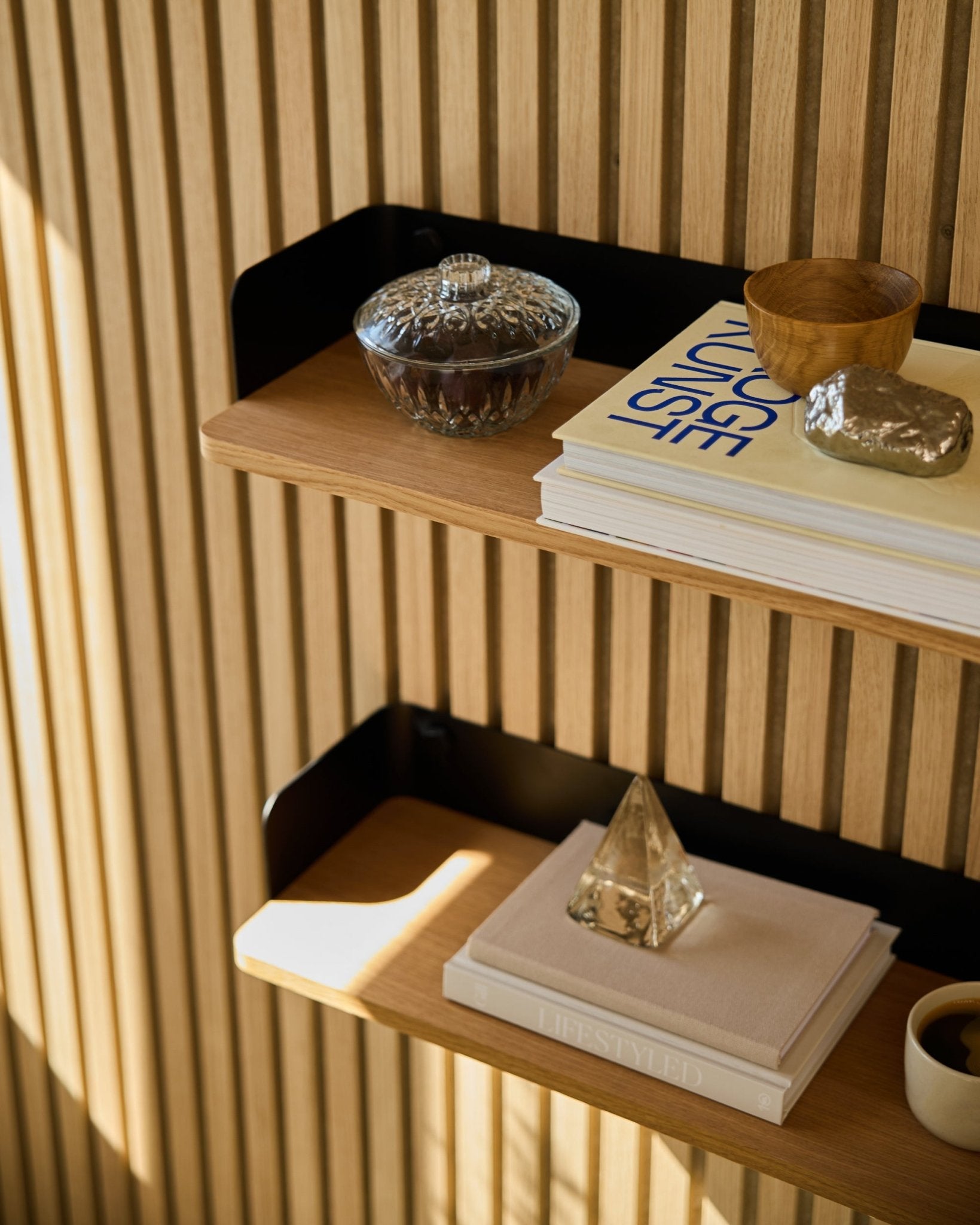 WoodUpp Linn Bücherregal in Natural Oak (55,9 cm) mit einem Glasgefäß, einer Tasse, einer goldenen Pyramide und Büchern vor Akupanel Akustik-Holzpaneelen im Sonnenlicht.