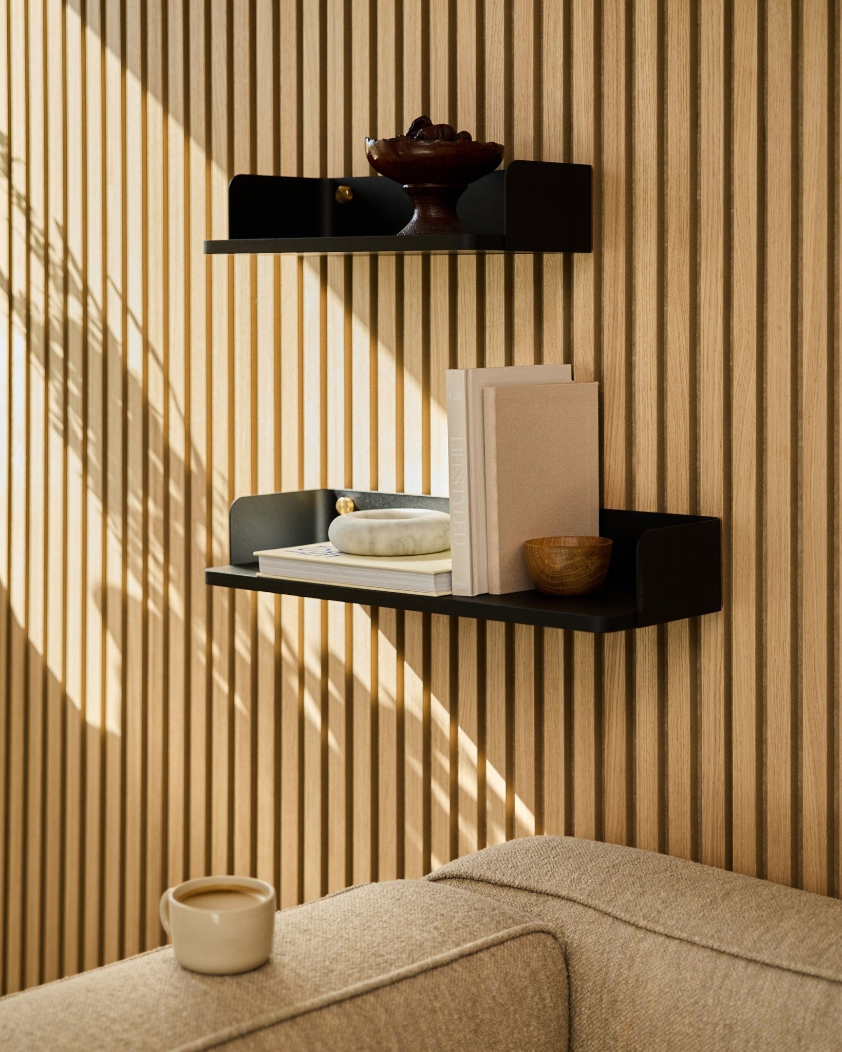 Ein black oak WoodUpp Bücherregal (55,9 cm) präsentiert Bücher, Dekor und eine Schale an einer Akustikholzpaneel-Wand; ein beige Sofa und eine Tasse stehen im Vordergrund.