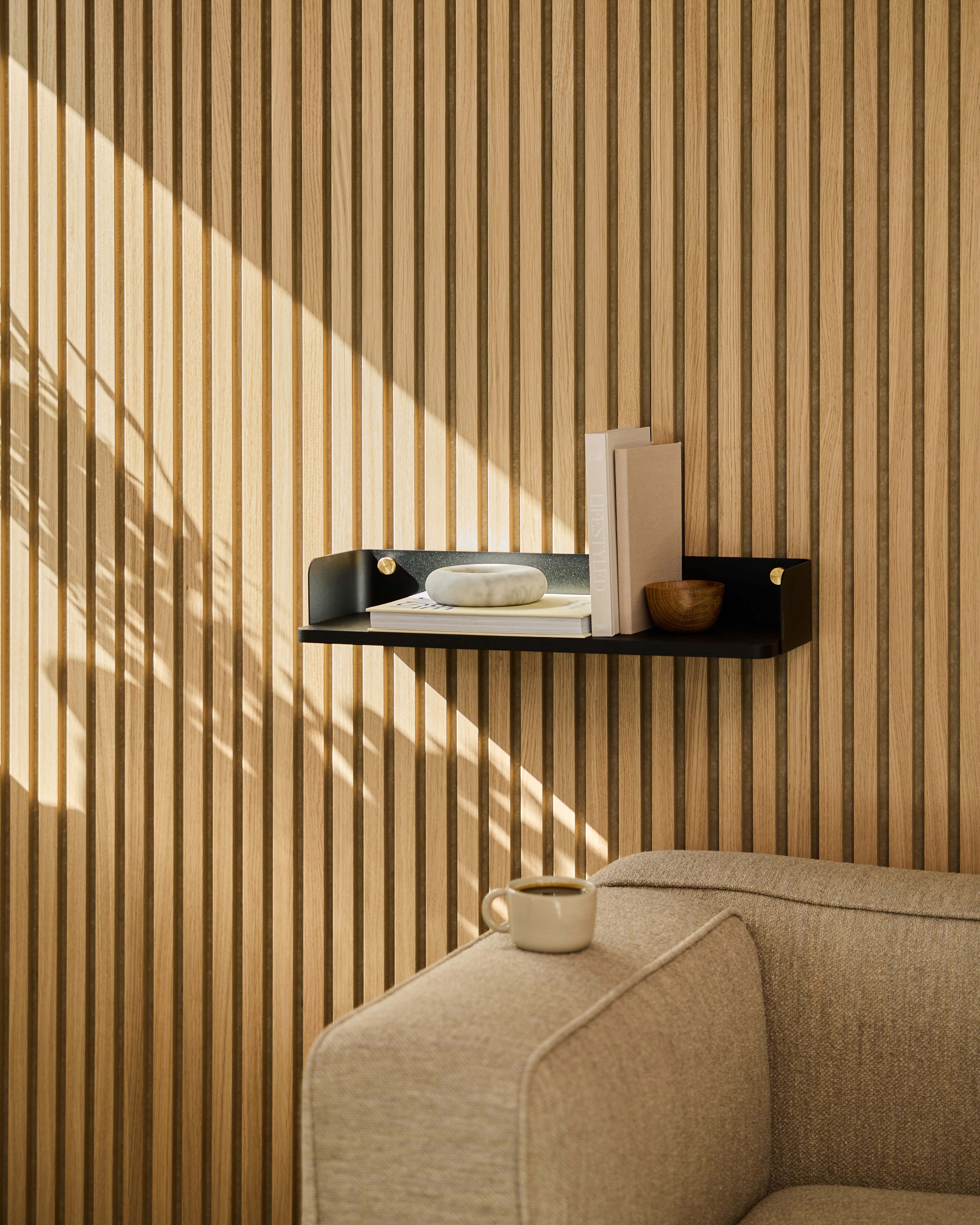 Ein Regal A Linn | Bookshelf / Black Oak von WoodUpp ziert die Wand über einem beigen Sofa. Das Sonnenlicht wirft Schatten auf die Wand aus gestreiften Akustikholzpaneelen.