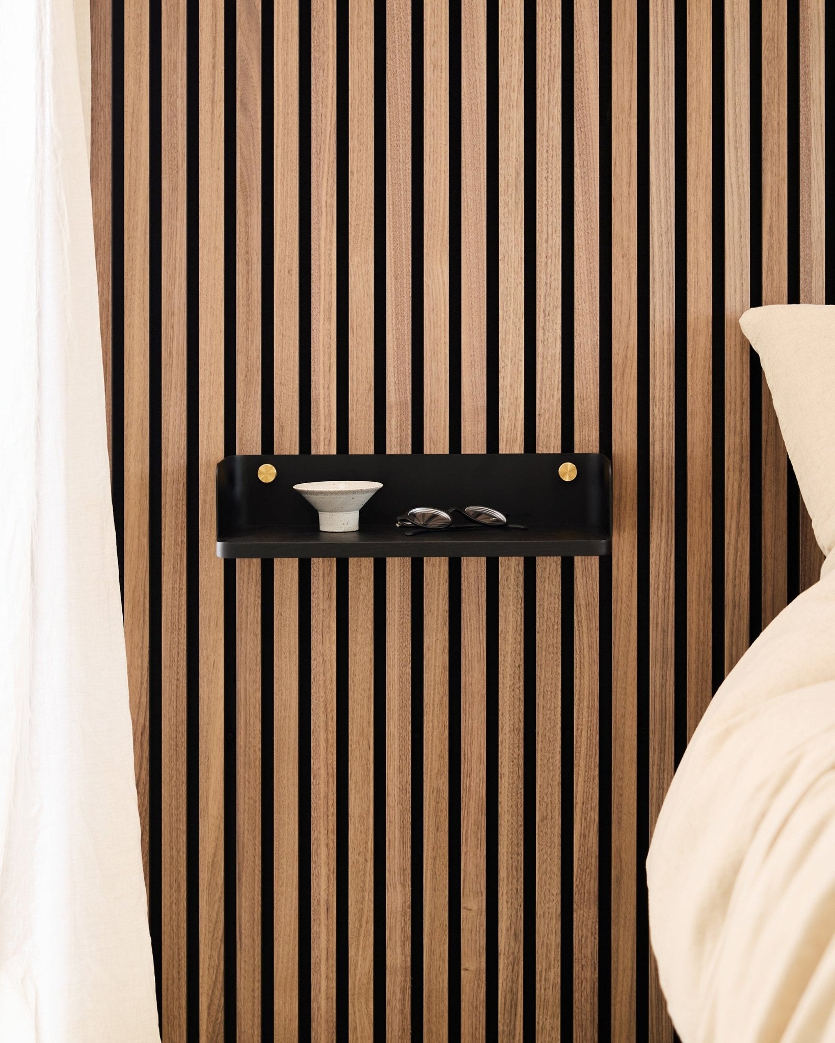 Das Linn Bücherregal in Black Oak von WoodUpp ist an einem nordischen Akustik-Lamellenpaneel montiert und trägt eine Schale und Gläser neben einem Bett mit Bettwäsche in beige.