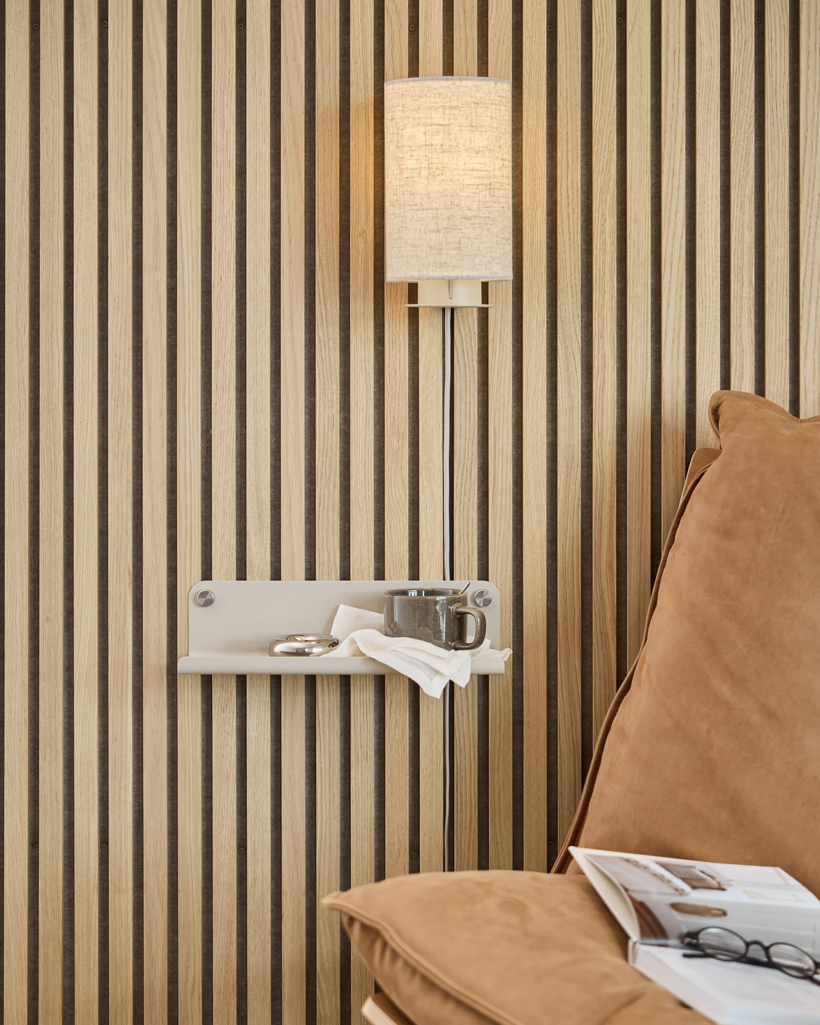 Vertikale Akupanel-Akustikwand aus Holzlamellen, beigefarbene Wandleuchte, kleines Regal mit Tasse und Serviette, hellbraunes Sofa mit offen liegender Zeitschrift.