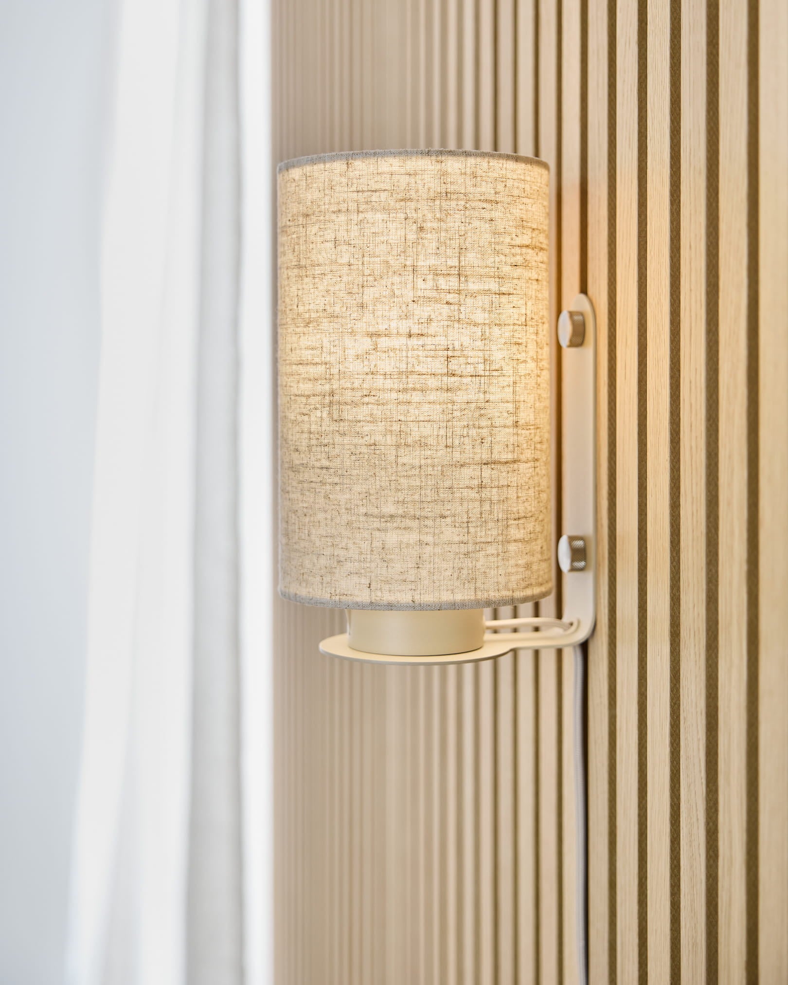 Die Feya Wandleuchte in Beige von WoodUpp wirft warmes Licht auf eine beige Akupanel Akustik-Lamellenwand und ist neben einem weißen Vorhang und einem Schirm aus Leinen-Baumwolle platziert.