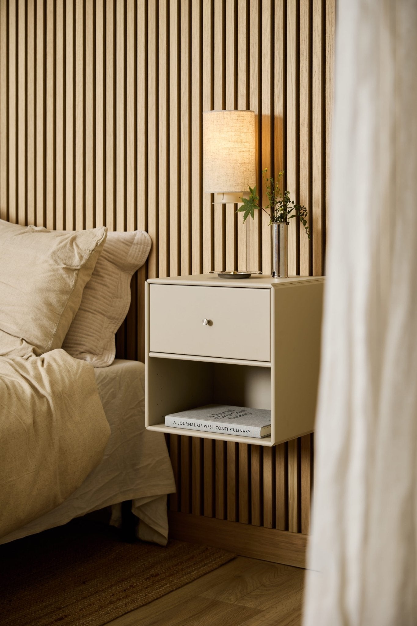 Auf einer beigen WoodUpp Arka-Bücherregalschublade neben dem Bett stehen eine Lampe, Bücher und eine Vase, vor dem Hintergrund einer WoodUpp Akupanel-Akustikwand aus Holzlamellen.