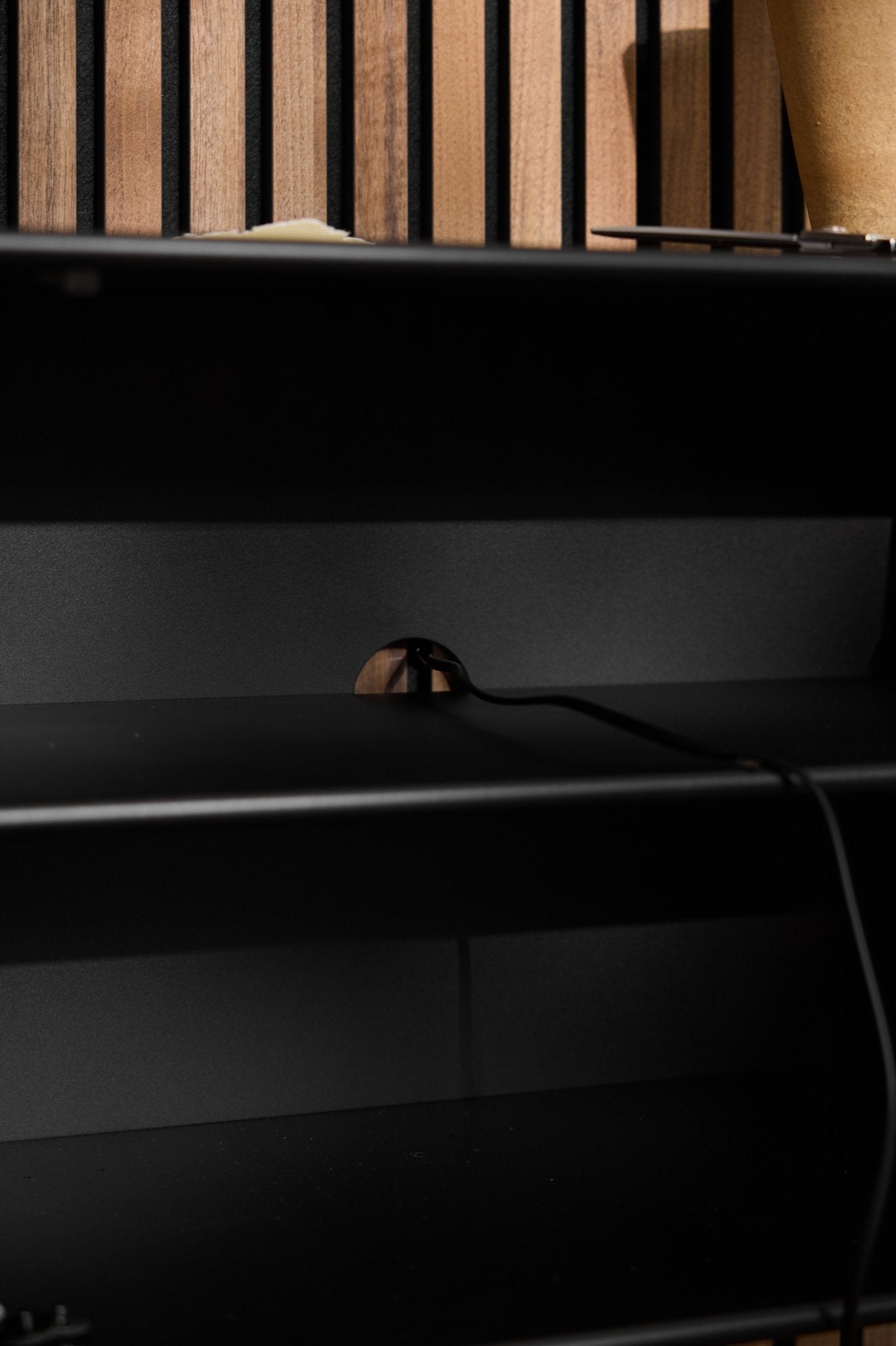 Schwarzer Arka-TV-Schrank von WoodUpp mit einer Kabeldurchführung in der Rückwand, aufgestellt vor einer vertikalen Akustikwand aus Holzlamellen.