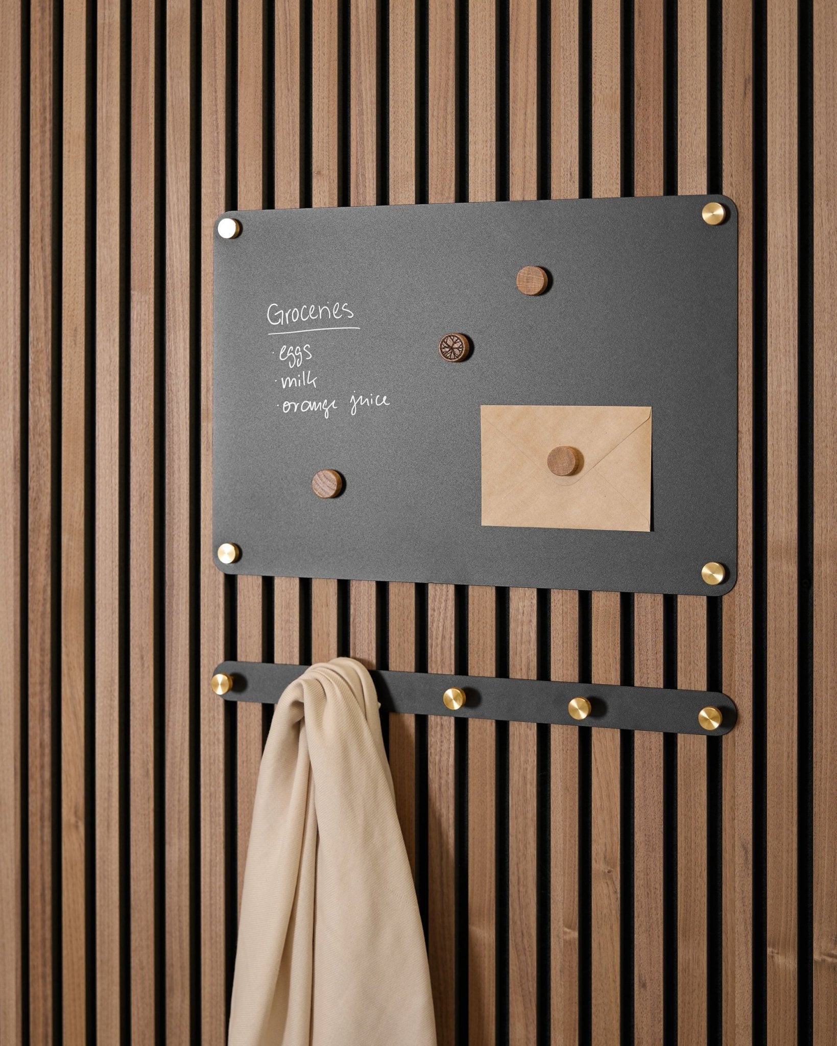 Eine small black Norr Magnettafel von WoodUpp trägt eine Einkaufsliste, einen Umschlag, Magnete und ein beige Tuch an Haken und ist an einem Akupanel Akustikpaneel montiert.