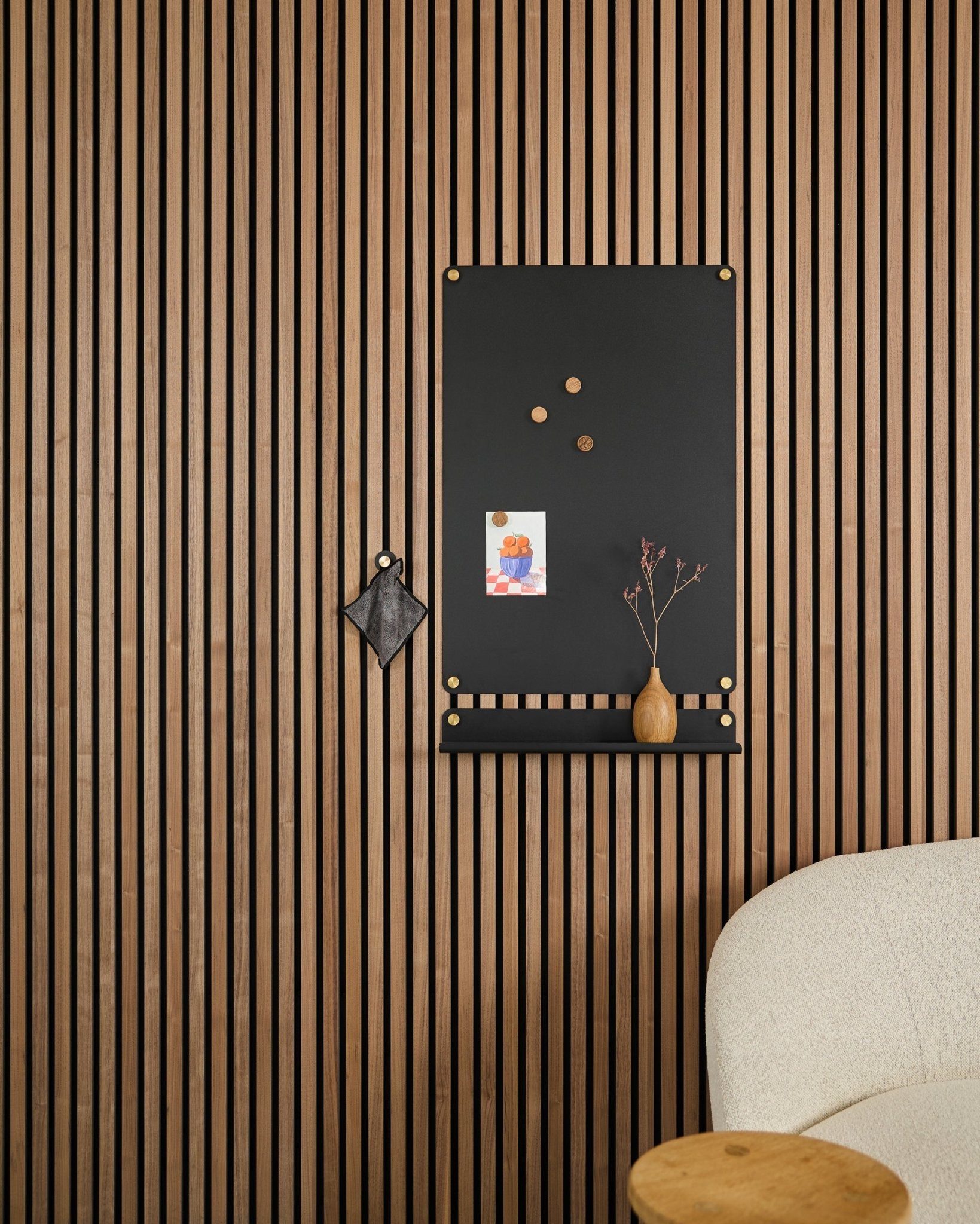 Eine large black Norr Magnettafel von WoodUpp, entwickelt für Akustikpaneele, hängt neben einem weiß gepolsterten Stuhl mit Notizen, einer Vase und einem kleinen Deko-Objekt.