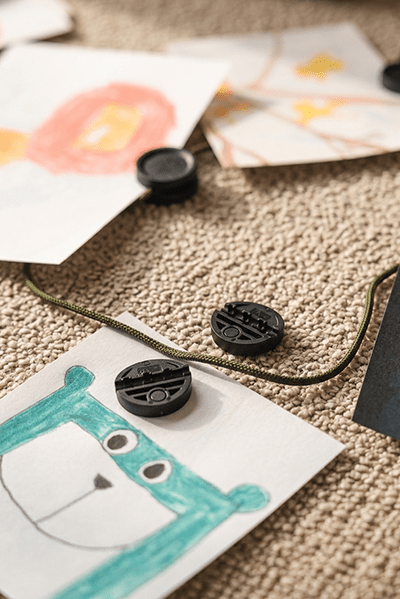 Kinderzeichnungen hängen mit Neodym-Clips an der WoodUpp Vindar | Gallery String; darunter liegt grüne Bärenkunst auf dem beige Teppich unter dem Akustikpaneel.