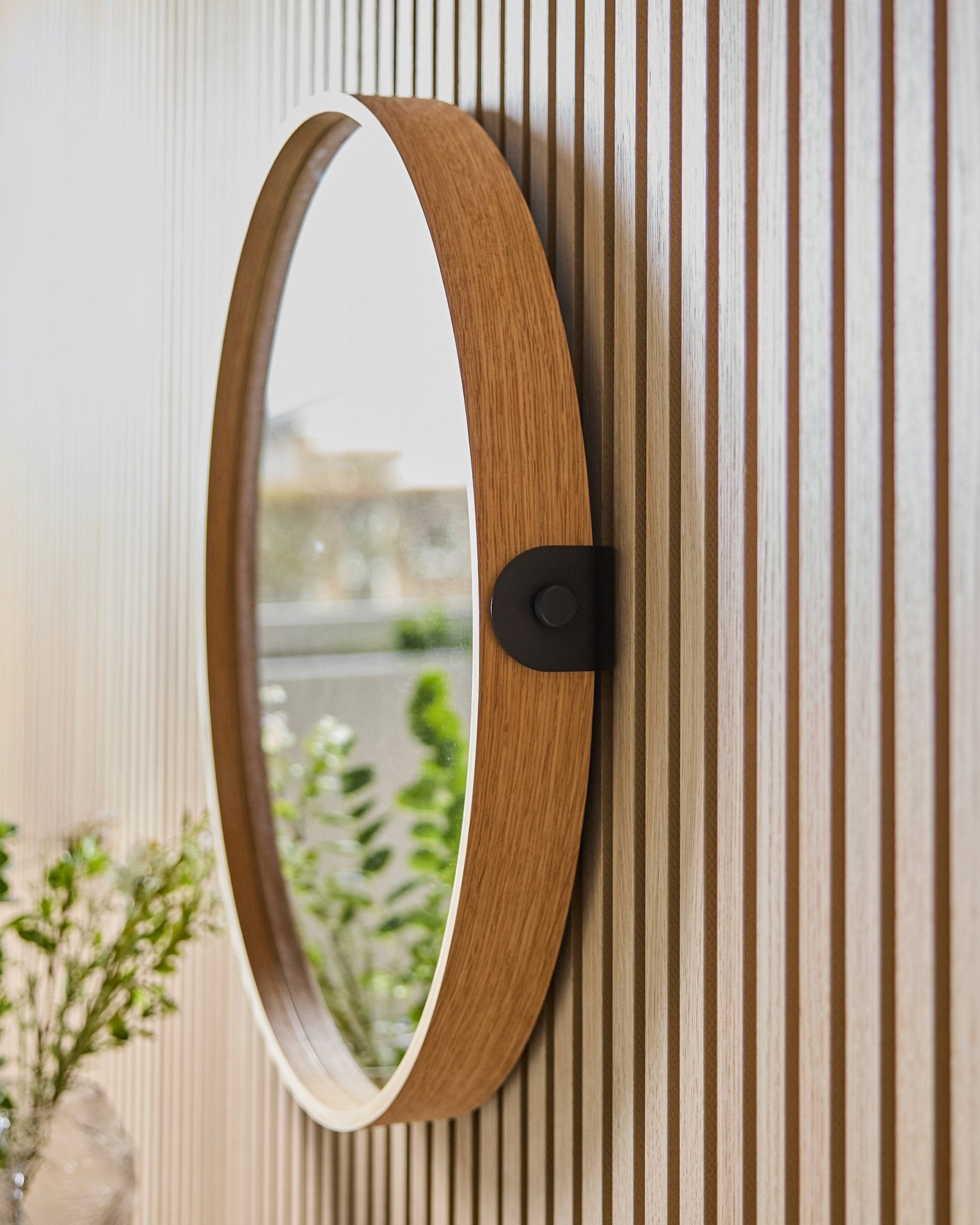 Linn | Mirror / Natural Oak von WoodUpp, montiert an einer gestreiften Wand aus Akustikpaneel, reflektiert Grünpflanzen und natürliches Licht aus einem nahen Fenster.