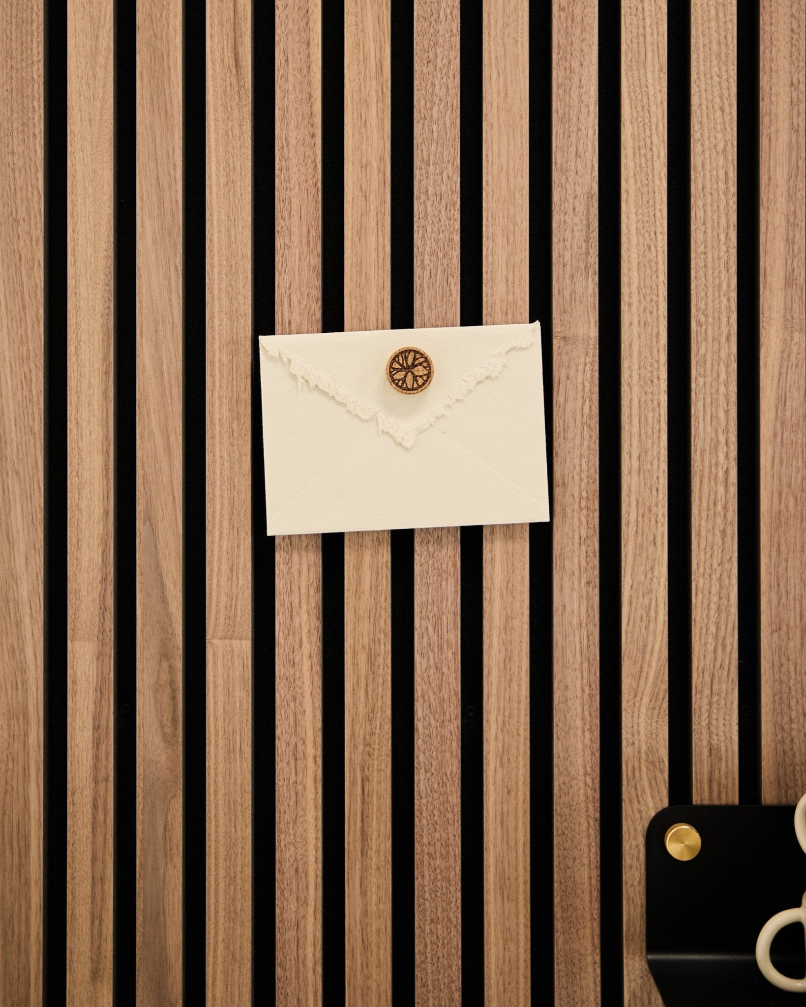 Ein cremefarbener Umschlag mit Wachssiegel ist mit dem Norr Poster Magnet-Set in Natural Oak an einem WoodUpp Akustik-Lamellenwandpaneel aus Eiche befestigt; Schere und Tablett liegen daneben.