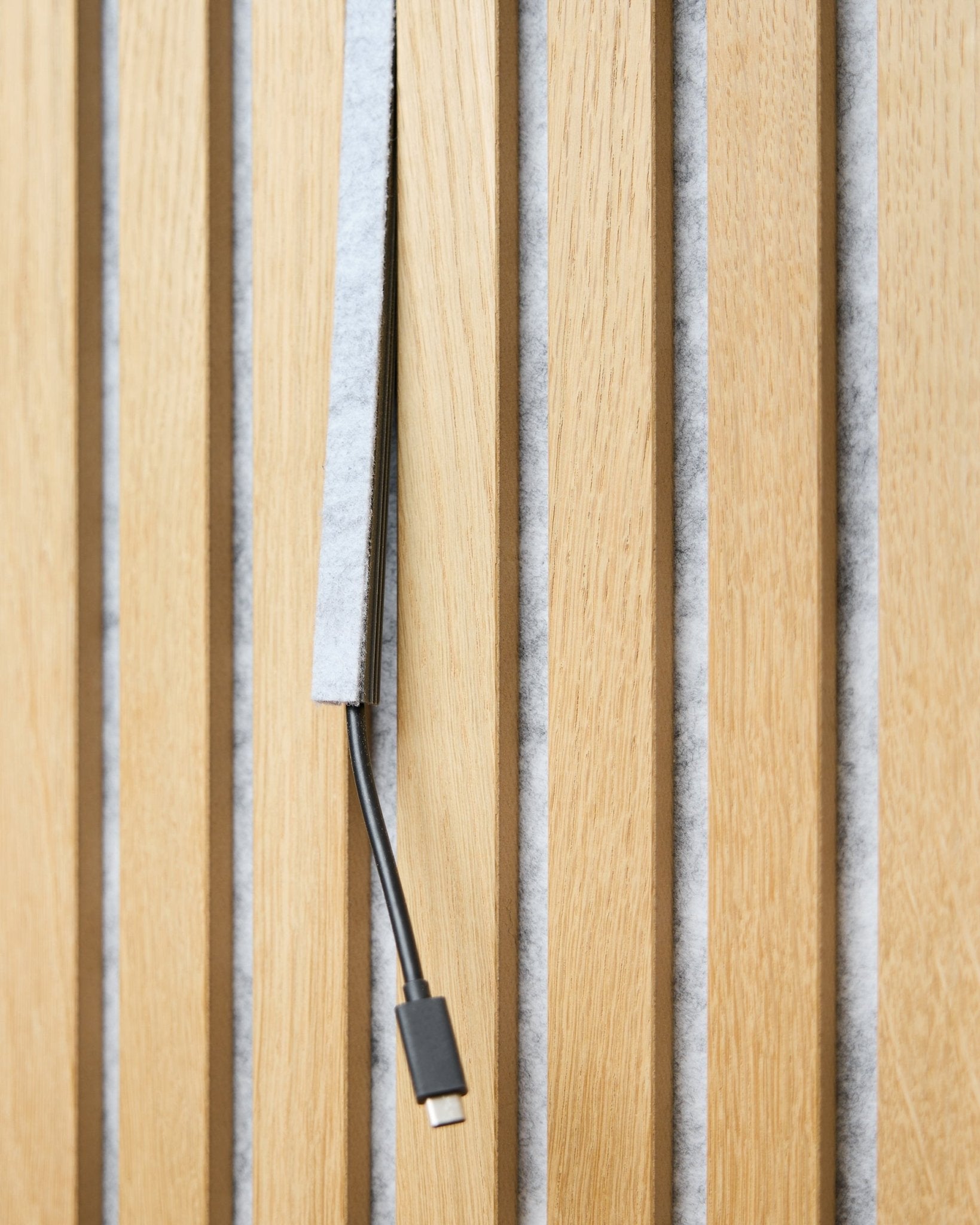 Ein black USB-C-Kabel verläuft durch eine grey WoodUpp Dupps Pop-on Cable Cover zwischen vertical akustischen Holzlamellen mit grey Filzrückseite.