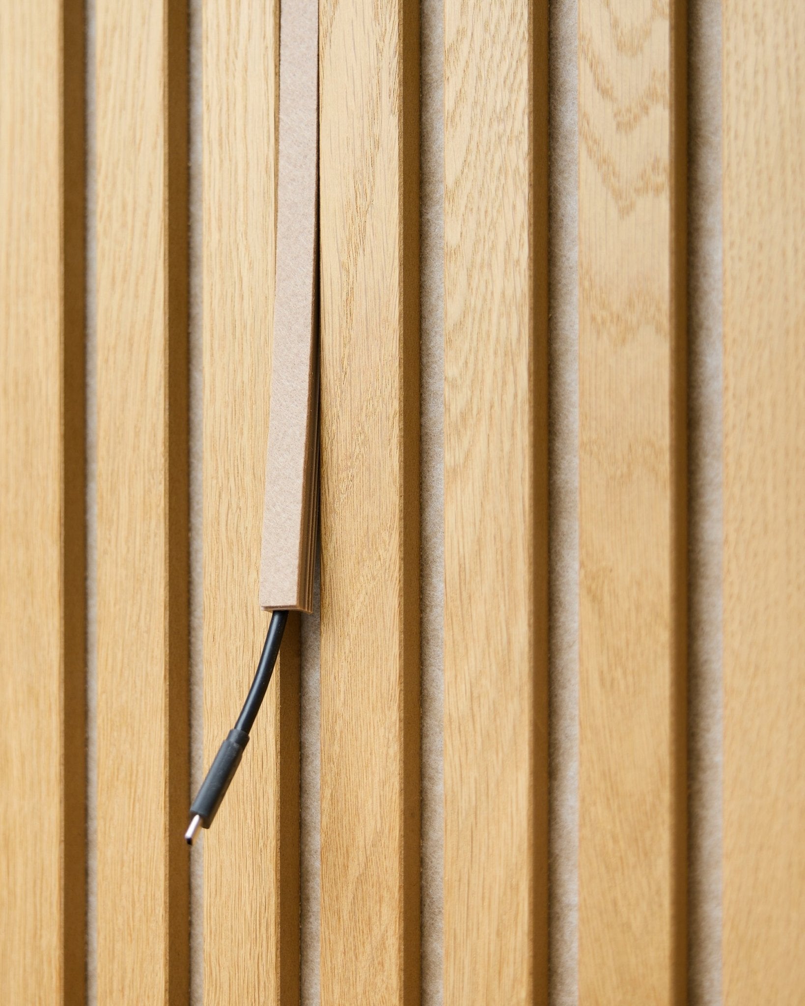 Ein black Kabel mit Kabelmanagement ist teilweise in einer WoodUpp Dupps Beige Pop-on Kabelabdeckung verborgen, montiert auf vertical Akupanel Akustik-Holzlamellen.