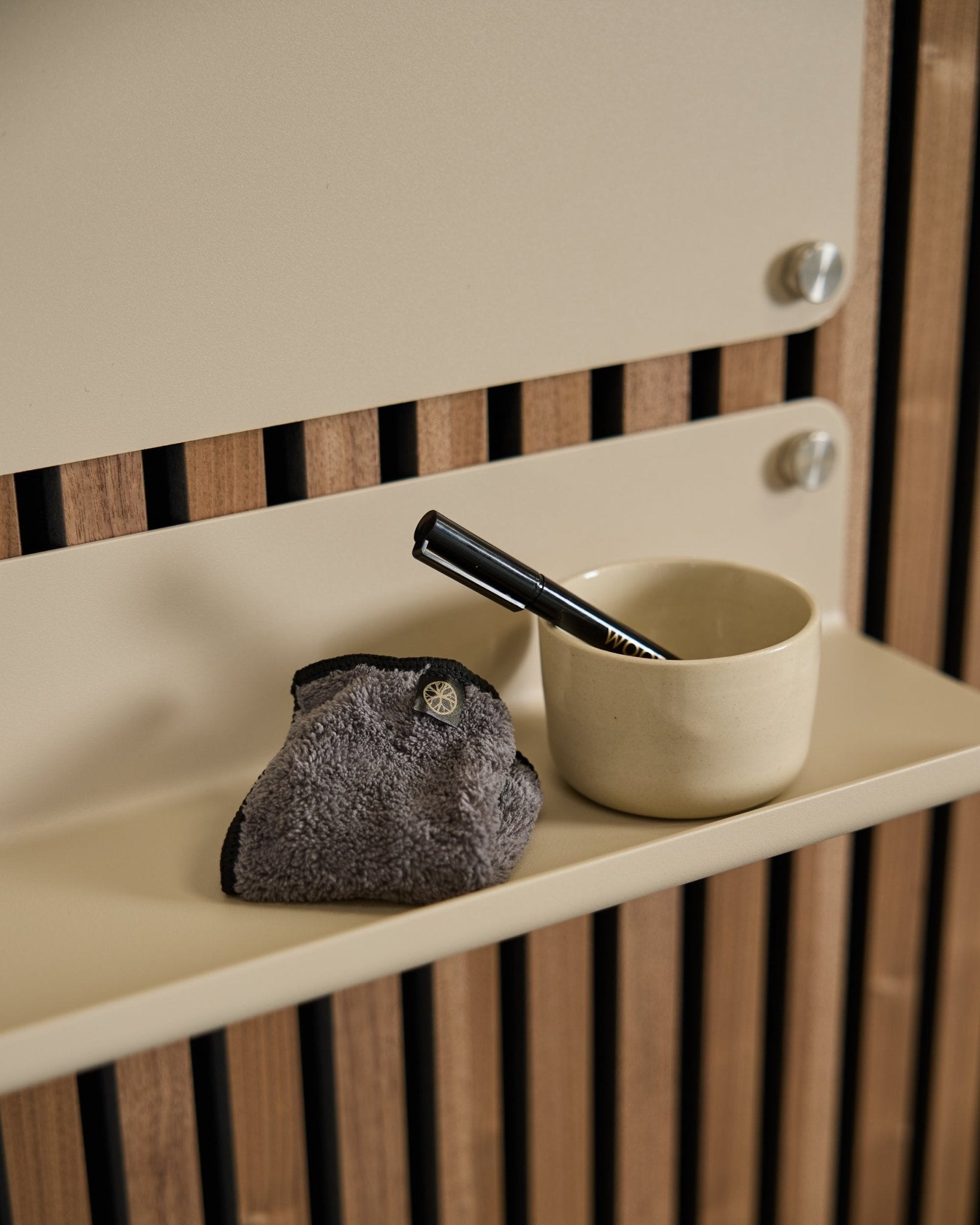 Das WoodUpp Norr Blackboard-Set umfasst eine small beige Tasse, eine Bürste mit black Griff und ein Reinigungstuch auf einem beige Regal vor nordischer Akustik-Holzverkleidung.