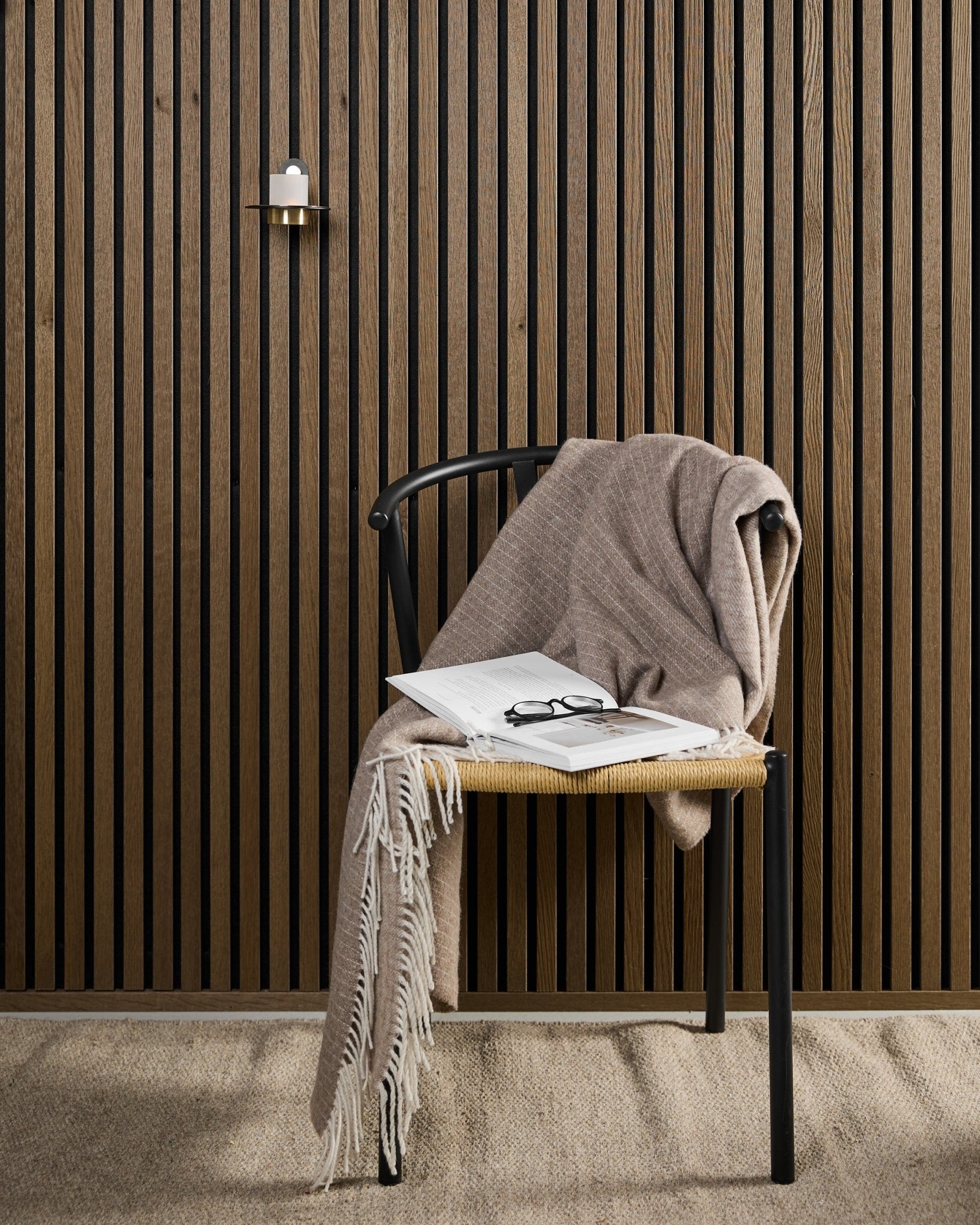Ein moderner Stuhl mit einer beige Wolldecke, einem offenen Buch und einer Brille steht neben einer Akupanel Akustik-Holzpaneele sowie einem black und brass WoodUpp Norr Kerzenhalter.