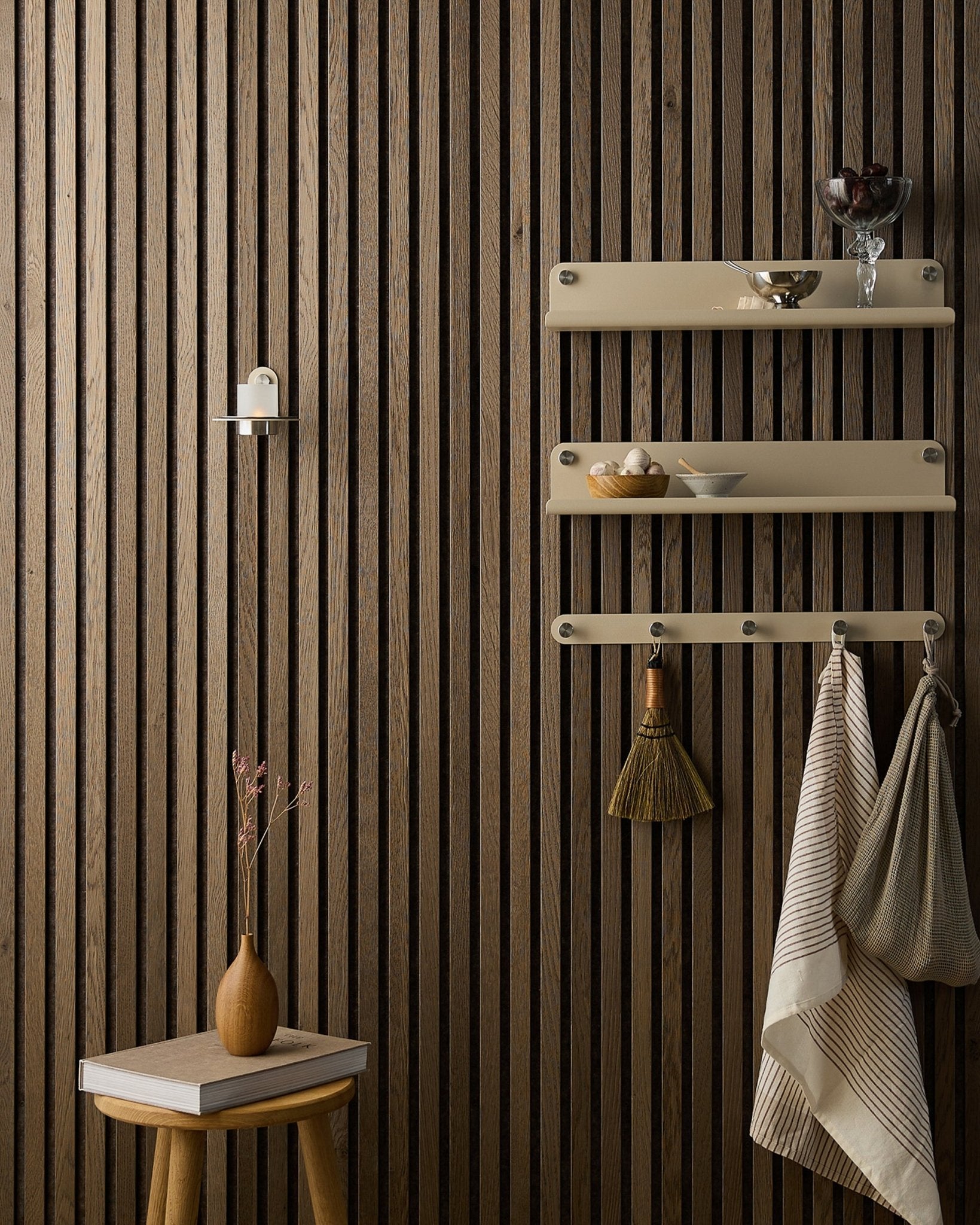 A wooden stool with dried flowers stands next to a WoodUpp Akupanel acoustic panel, beside beige shelves holding dishes, a brush, towels, and a Norr Candle Holder.