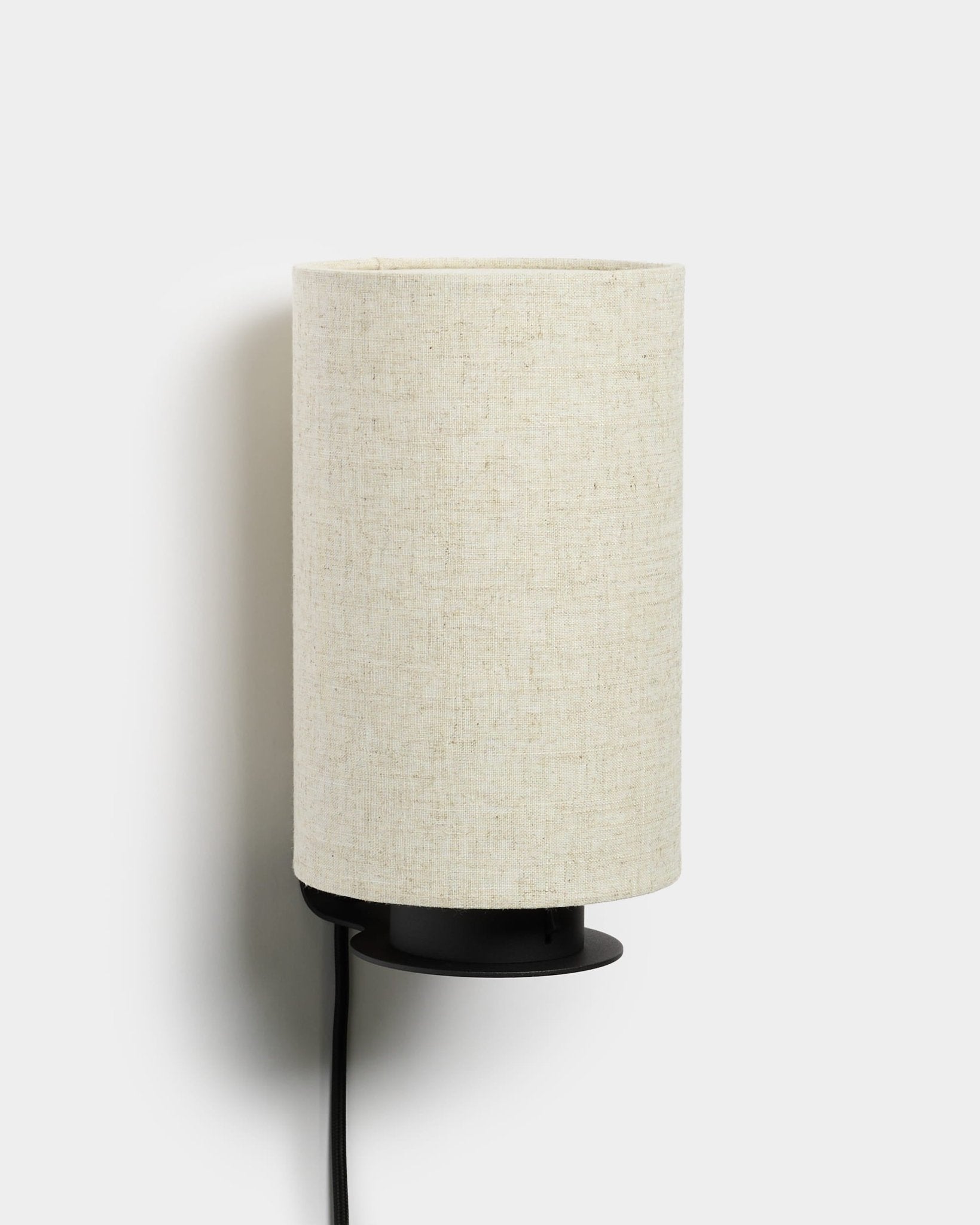 Feya Wandleuchte von WoodUpp mit einem Metallsockel in black, einem strukturierten Leinenschirm aus Baumwollmischung in beige und einem sichtbaren Stromkabel in black. Nordisch-minimalistisches Design.