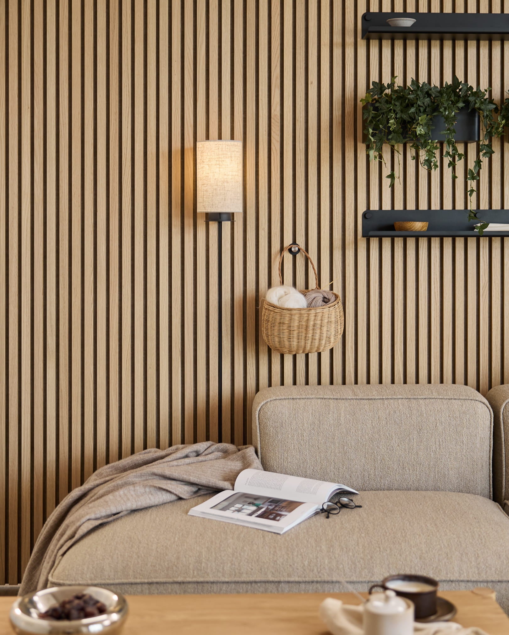 Sofa in Beige mit Decke und Buch neben einer WoodUpp Akustik-Lamellenwand in Akupanel, Regalen mit Pflanzen und Deko sowie einer Feya Wandleuchte in Black/Beige.