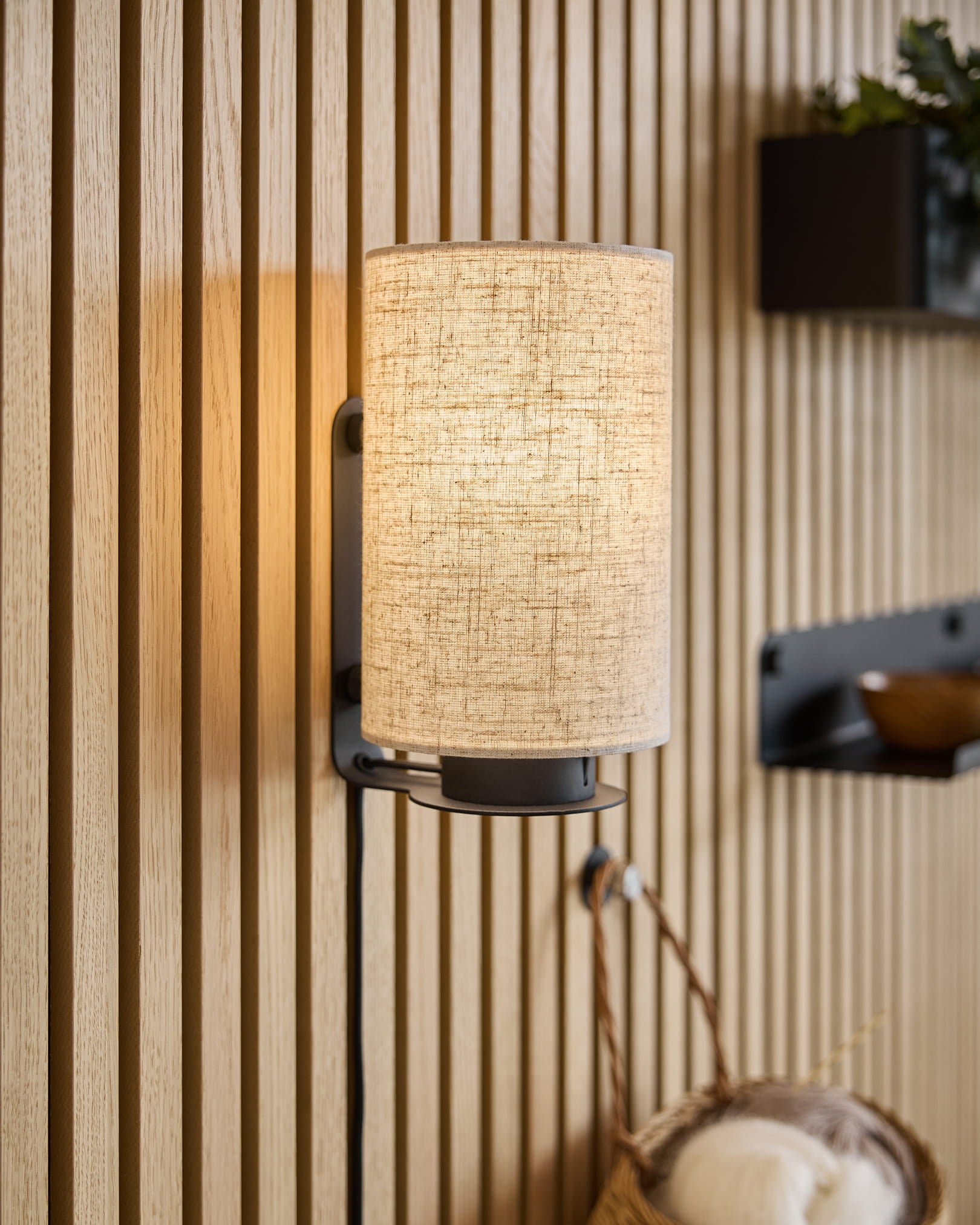 Ein moderner Wandleuchter mit Stoffschirm wirft warmes Licht auf Akupanel-Akustikholzplatten, die durch minimalistische Regale und einen geflochtenen Korb ergänzt werden.