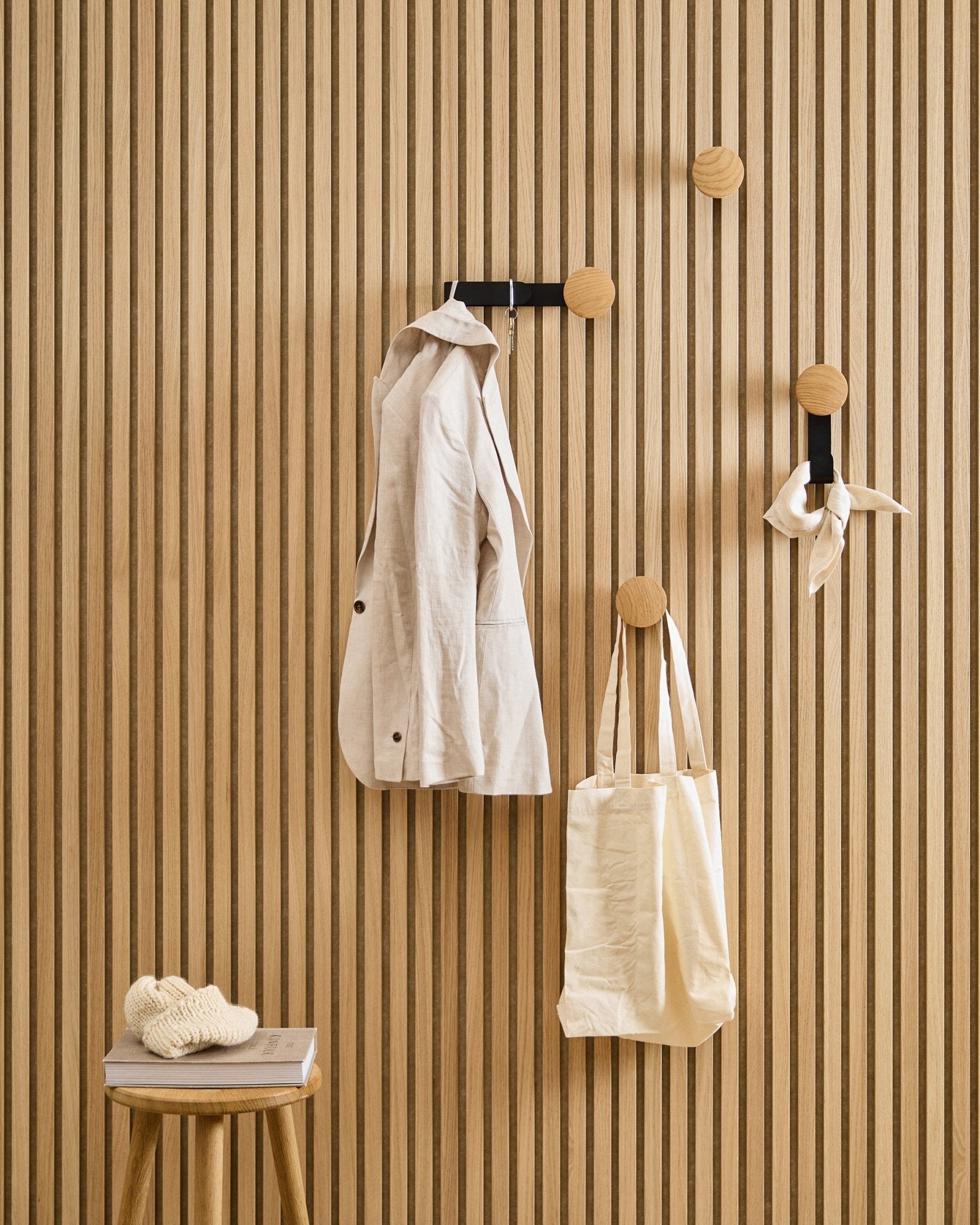 Eine beige Jacke, eine Tote Bag und ein Schal hängen am Linn Horizontal Haken von WoodUpp in Natural Oak; auf einem Hocker daneben liegen ein Buch und ein Strickstück.