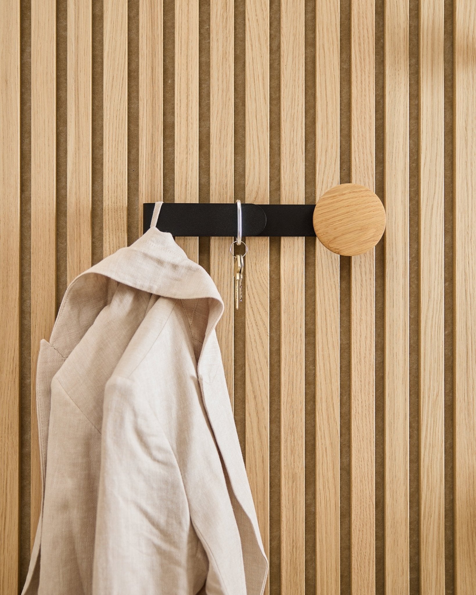 Ein beige Blazer und ein Schlüsselbund hängen am WoodUpp Linn Horizontal Haken in natural oak, montiert auf einem natural oak Akupanel Akustik-Holzpaneel.