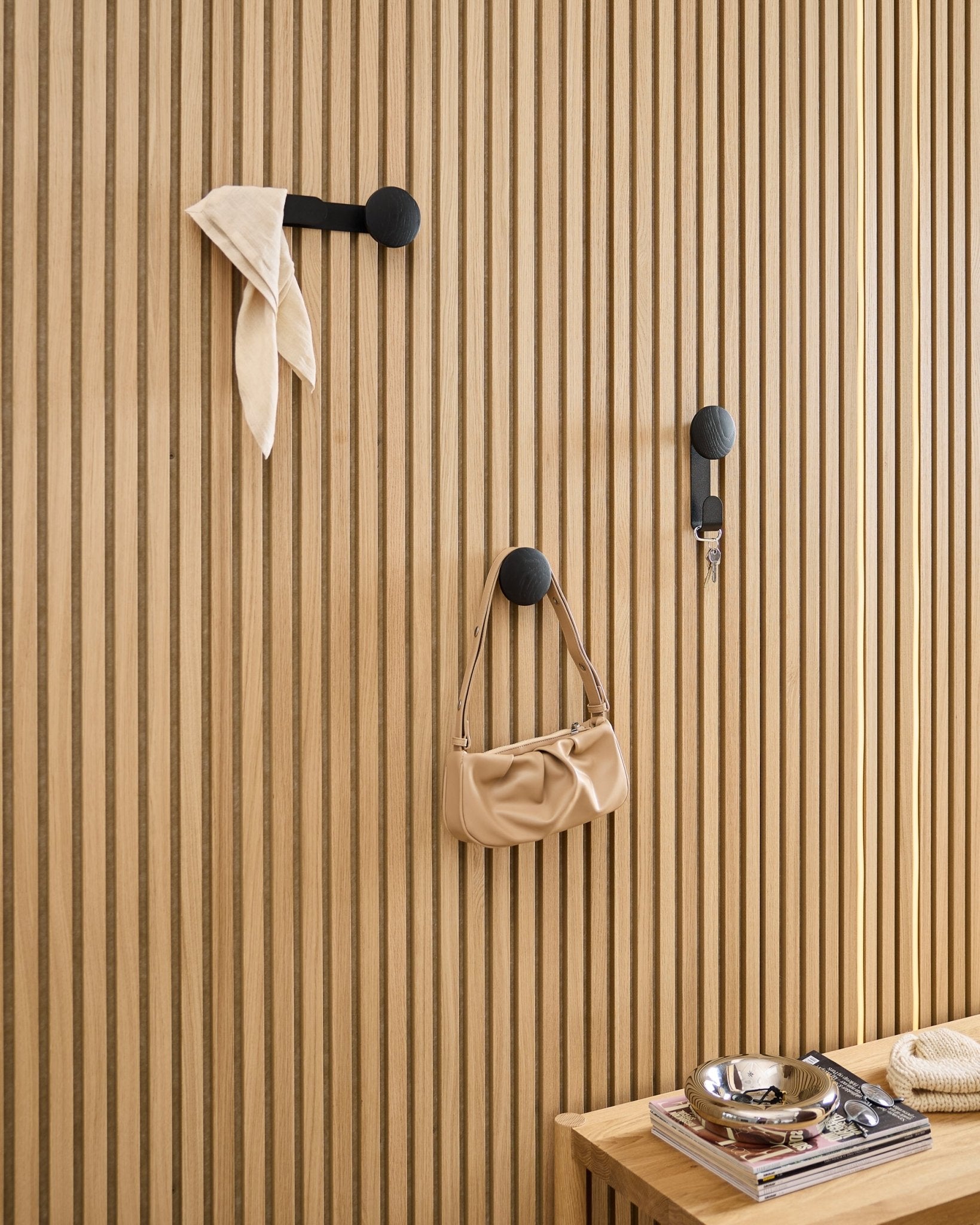 WoodUpp Linn Single Hanger in Black Oak trägt einen Schal, eine beige Tasche und Schlüssel an einem black Akupanel Akustik-Holzpaneel; daneben eine Bank mit Büchern und einer Metallschale.