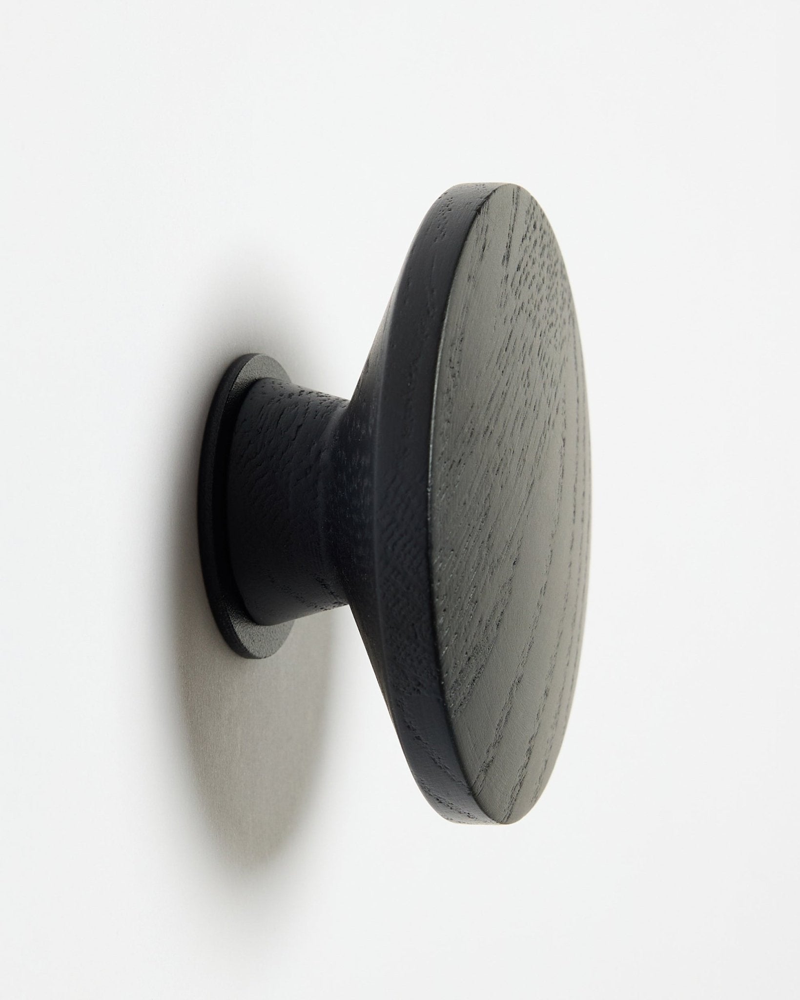 Ein rundes WoodUpp Linn | Single Hanger in black oak, entworfen als Accessoire für Akupanel Akustik-Holzpaneele, montiert auf einer weißen Oberfläche mit weichem Schatten auf der linken Seite.
