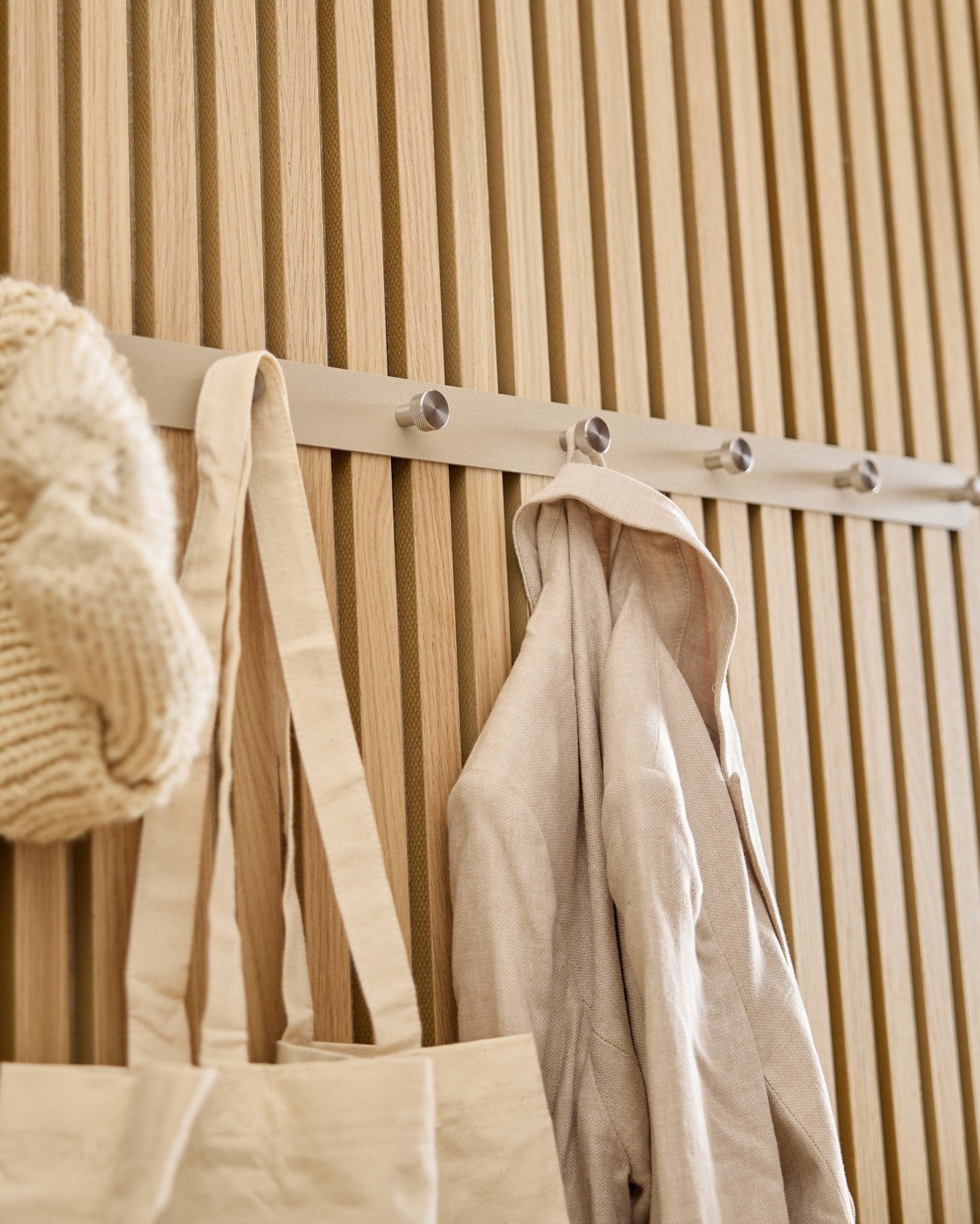 Eine beige Tote Bag, Strickmütze und ein Leinenhemd hängen an der WoodUpp Norr Garderobenhakenleiste (Beige/Edelstahl Steel, X7), montiert an einer hellen Holz-Lamellenwand in Akupanel.