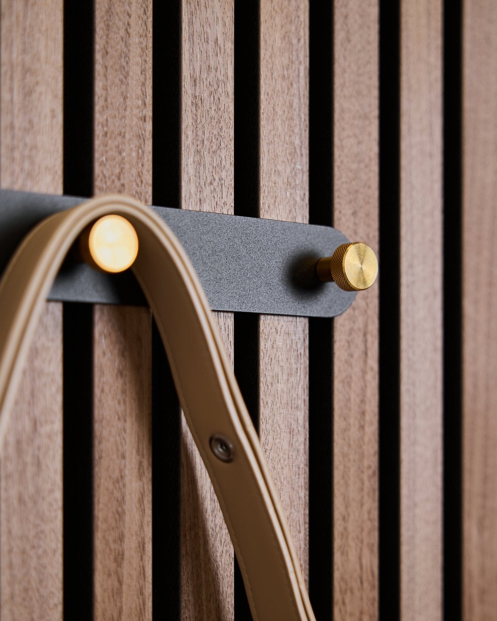 Fünf black/brass Haken der Norr Garderobenleiste von WoodUpp sind an einem Akustikpaneel mit Holzlamellen montiert und halten jeweils eine beige Schlaufe.