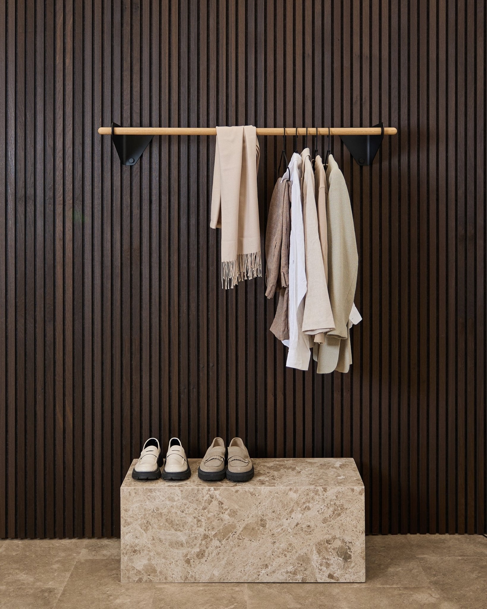 Kleidung in neutralen Tönen und ein Schal hängen an einer 115,9 cm langen Linn | Coat Rail in black von WoodUpp über einer Marmorbank, vor einem dunklen Akustikpaneel mit Lamellen in Akupanel.