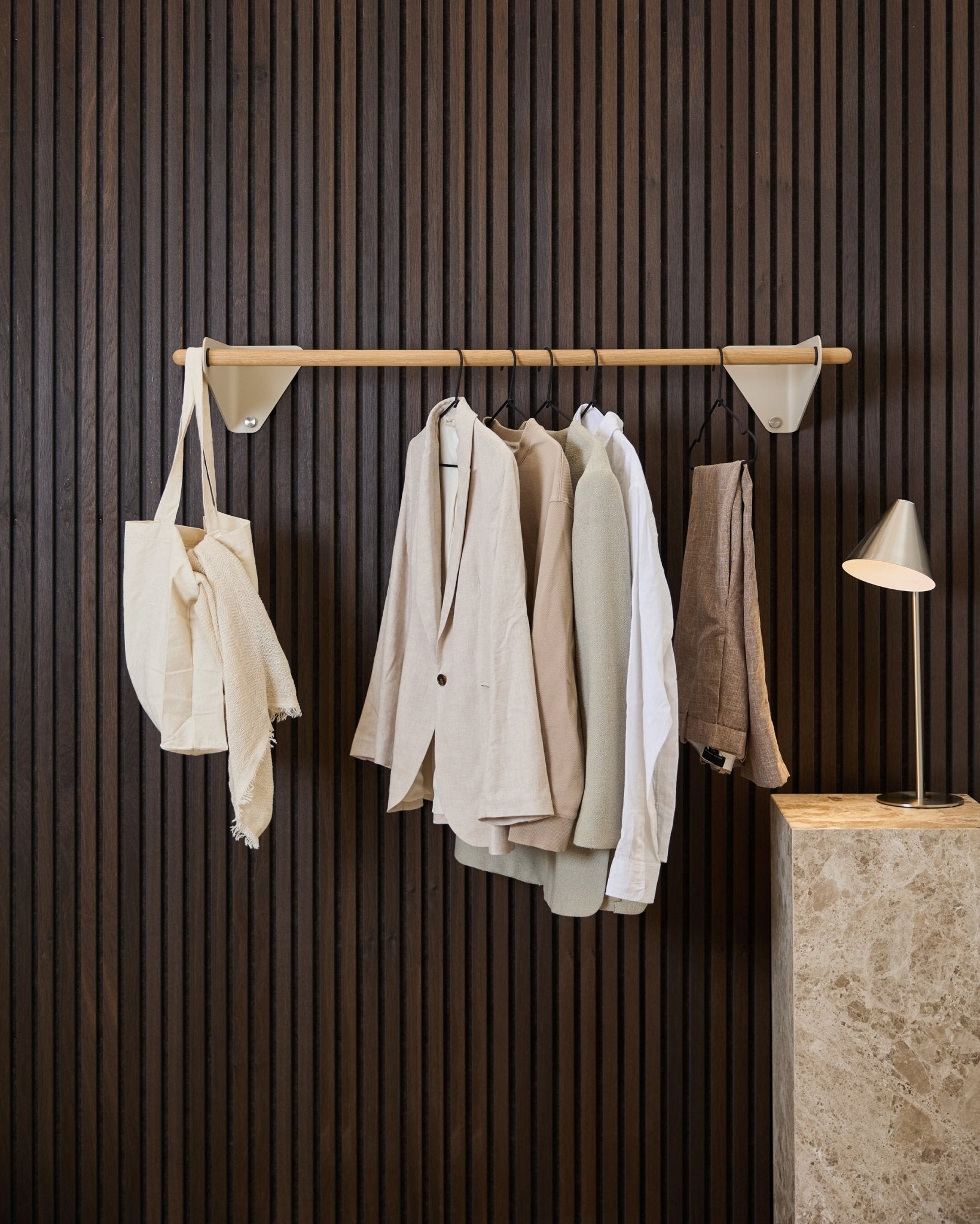 Eine beige WoodUpp Linn Garderobenstange (115,9 cm) trägt Jacken, ein weißes Hemd und eine cremefarbene Stofftasche vor einem dunklen Holz-Akupanel Akustikpaneel und einer Marmorlampе.