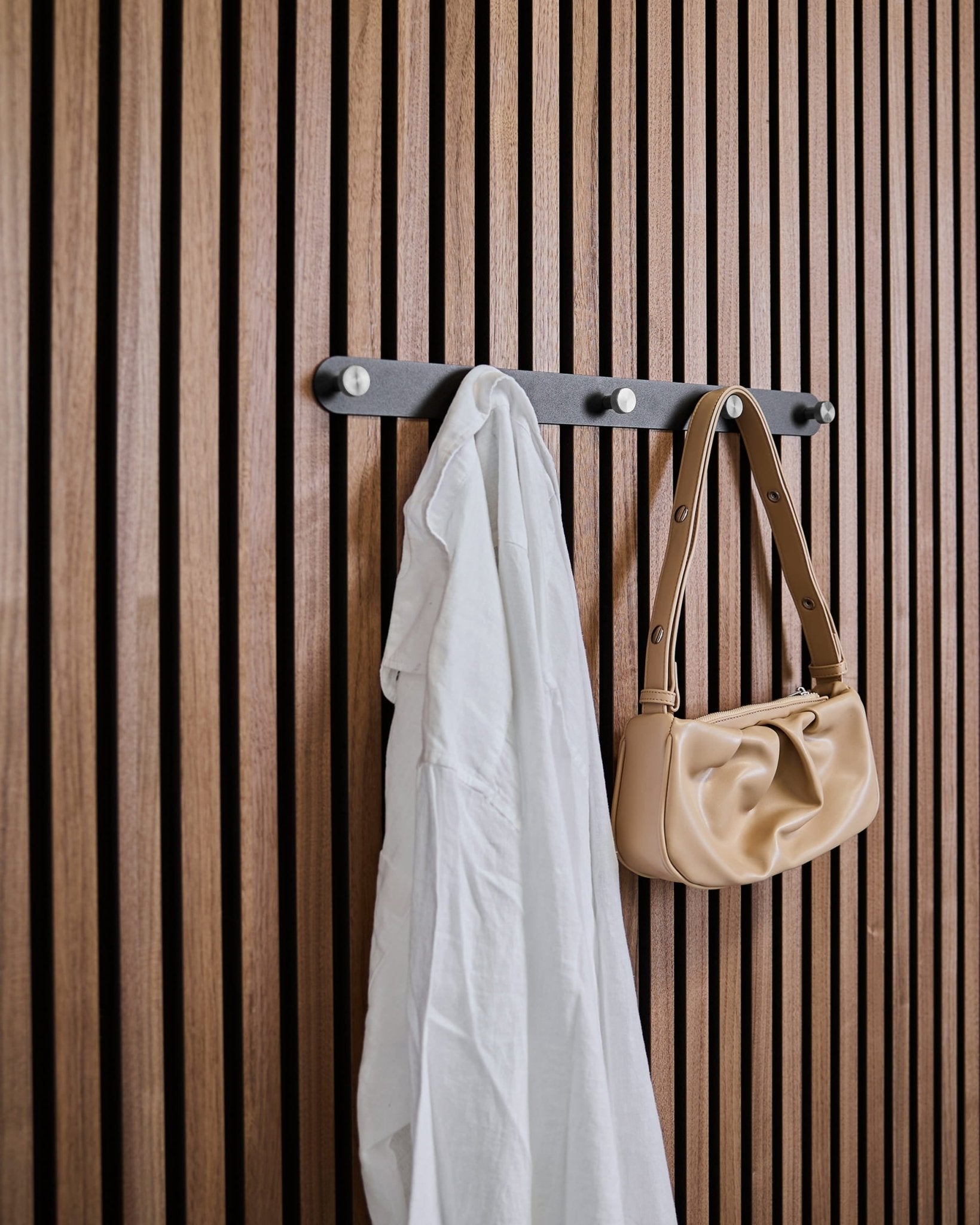 Ein weißes Hemd und eine beige Handtasche hängen an den WoodUpp Norr X5 Black/Stainless Steel Garderobenhaken, die an einem vertical Akustikpaneel mit Lamellen befestigt sind.
