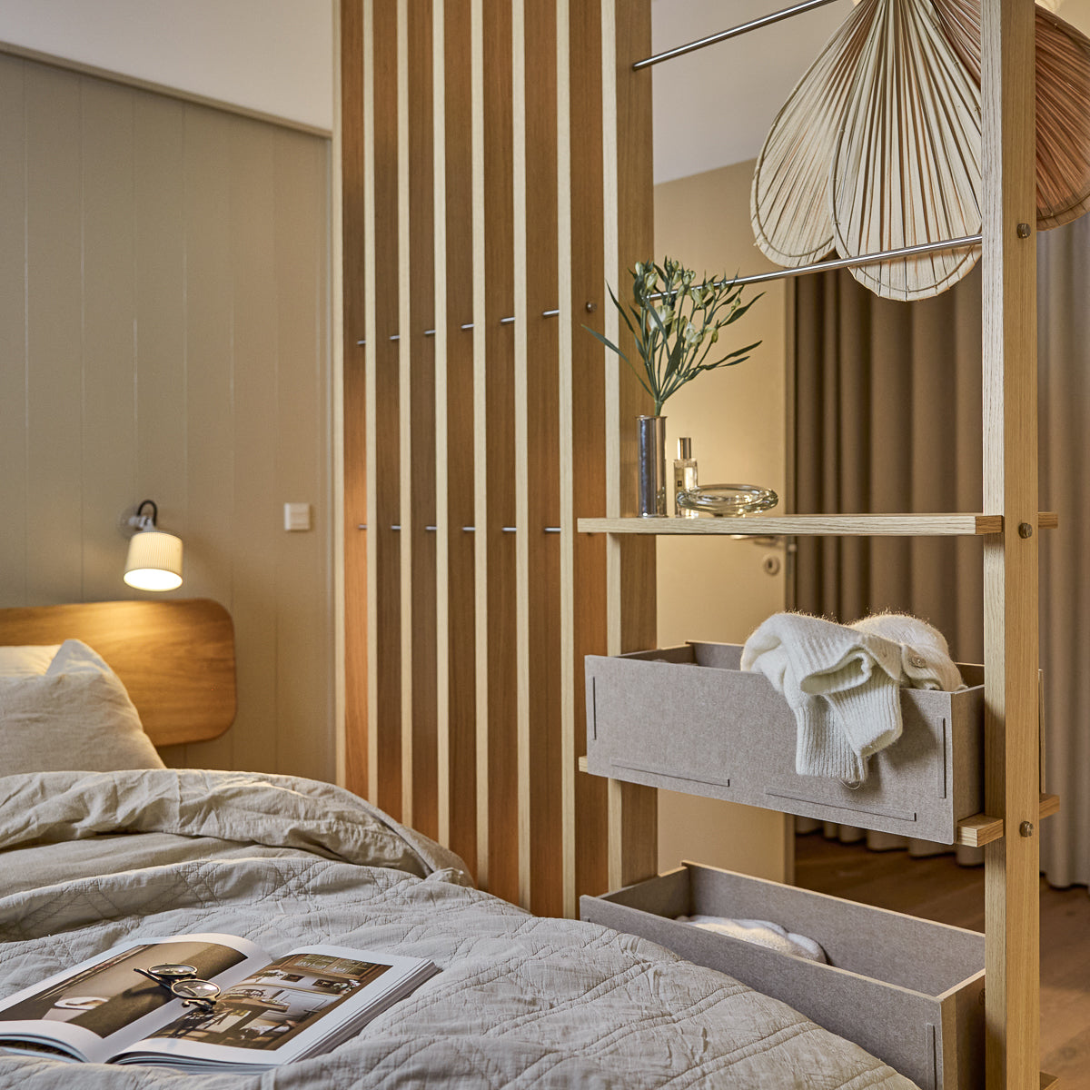 Ein modernes Schlafzimmer mit nordisch weicher Bettwäsche, Hängeleuchte und einer Zeitschrift auf dem ordentlich gemachten Bett. WoodUpp Akupanel Akustikholzplatten akzentuieren die Wand.