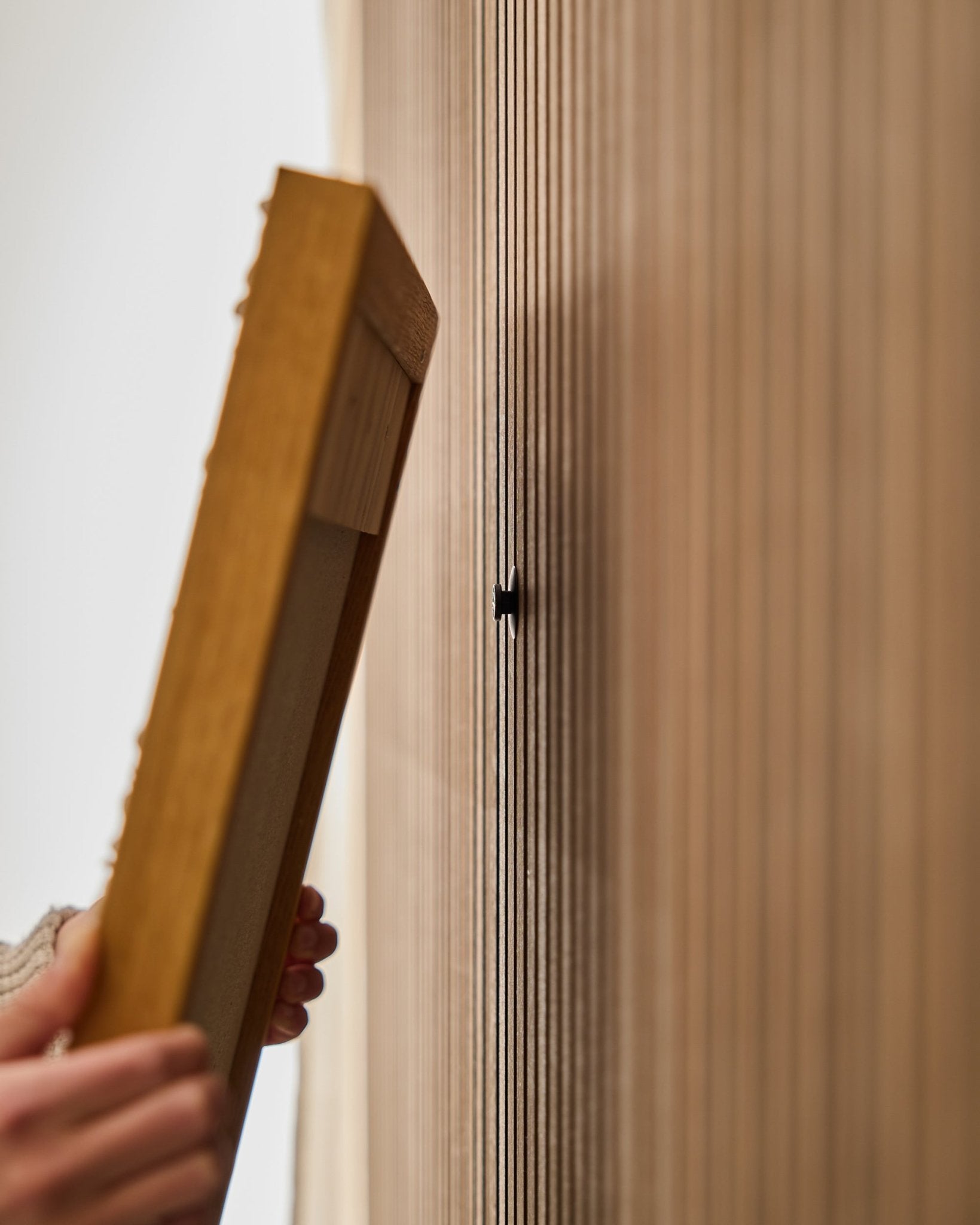 Eine Person nutzt das WoodUpp Dupps | Original Set, um einen Rahmen für ein Akupanel Akustik-Holzpaneel zu montieren, mit Fokus auf dem Anbringen eines small black Hakens nahe vertical Lamellen.