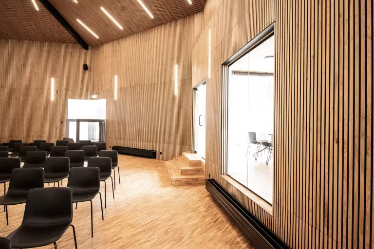 Geräumiger Konferenzraum mit Akupanel-Akustikholzplatten an Wänden und Decke, schwarzen Stuhlreihen, großen Fenstern und vertikalen Beleuchtungskörpern.