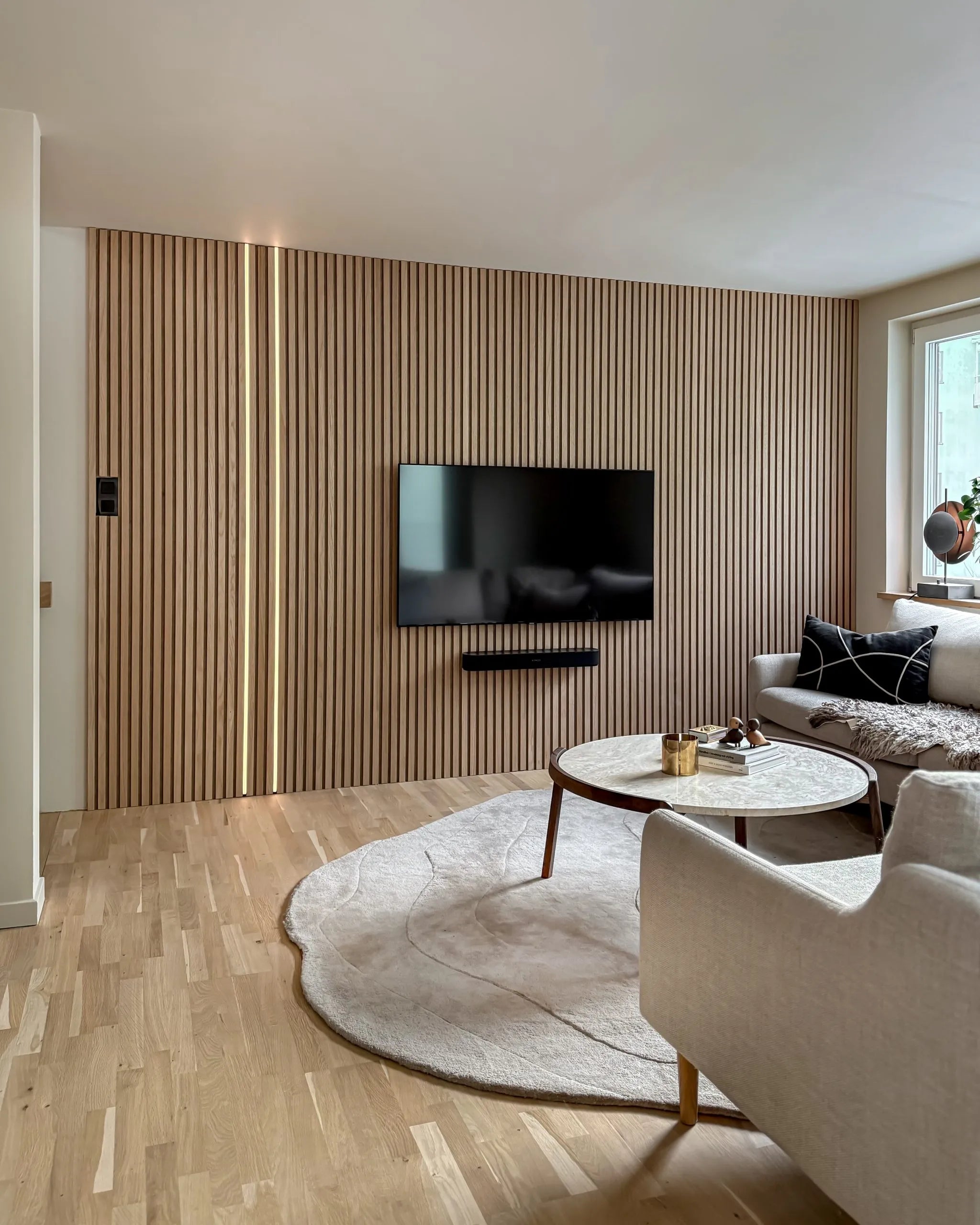 Modernes Wohnzimmer mit einer Akzentwand aus Akupanel-Akustikholz, montiertem Fernseher, rundem Couchtisch, cremefarbenem Sofa, großem Teppich auf hellem Holzboden.