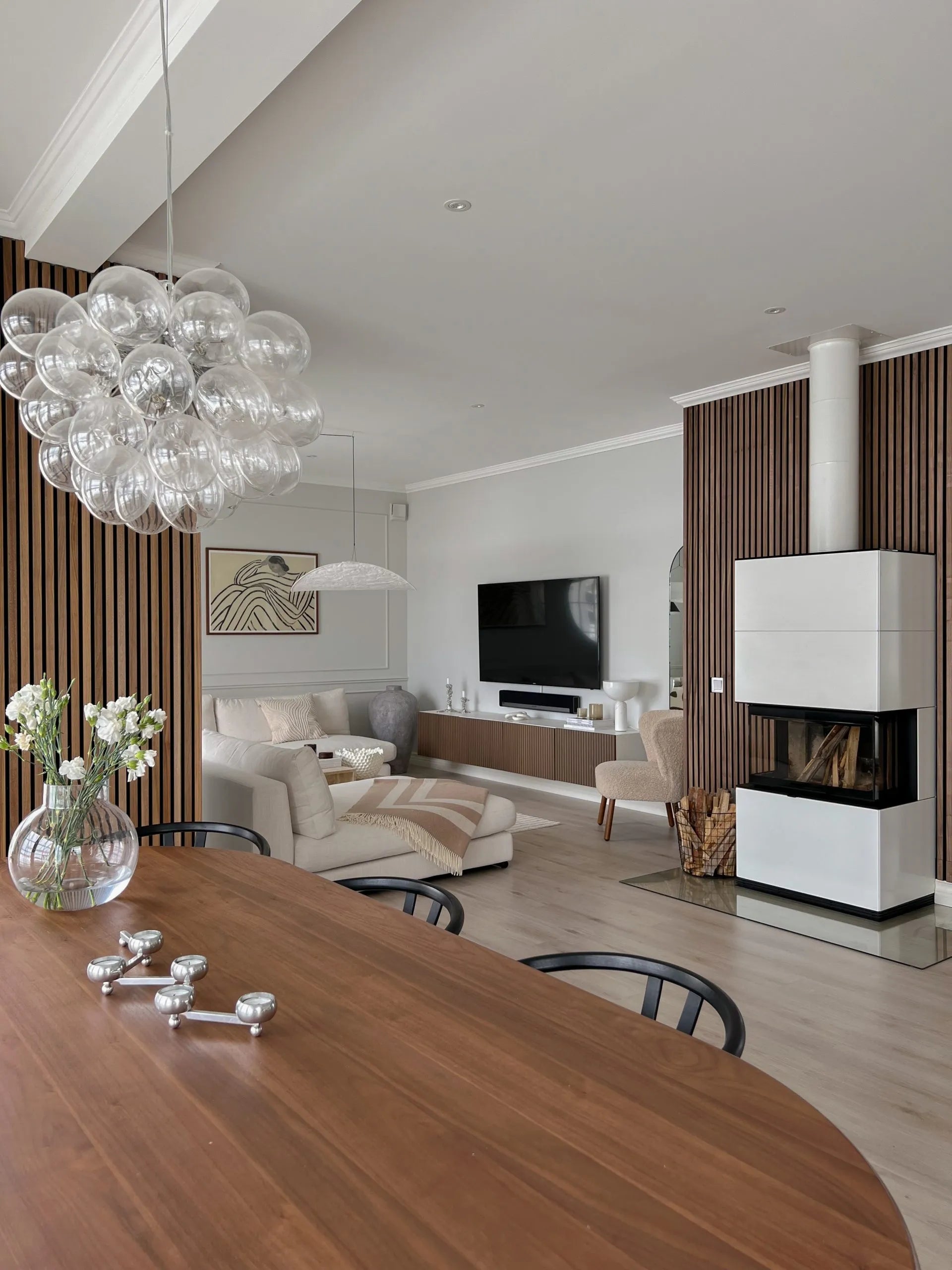 Moderner Wohn- und Essbereich mit Akustikpaneelen aus nordischem Holz, gemütlichen Sitzgelegenheiten, einem Fernseher, einem eleganten Kamin mit gestapeltem Brennholz und einem Kronleuchter mit Glaskugeln.