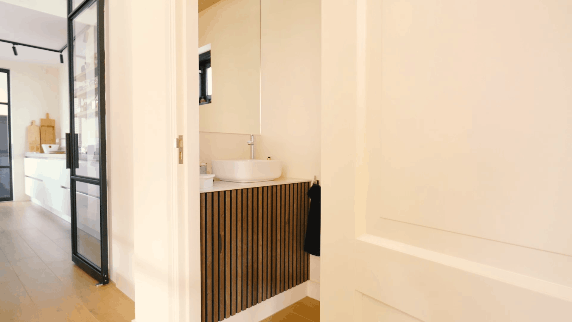 Modernes Badezimmer mit rundem Waschbecken, akustischem Waschtisch mit Holzlatten, großem Spiegel und weißer Tür neben einem hellen Flur und einer modernen Küche.