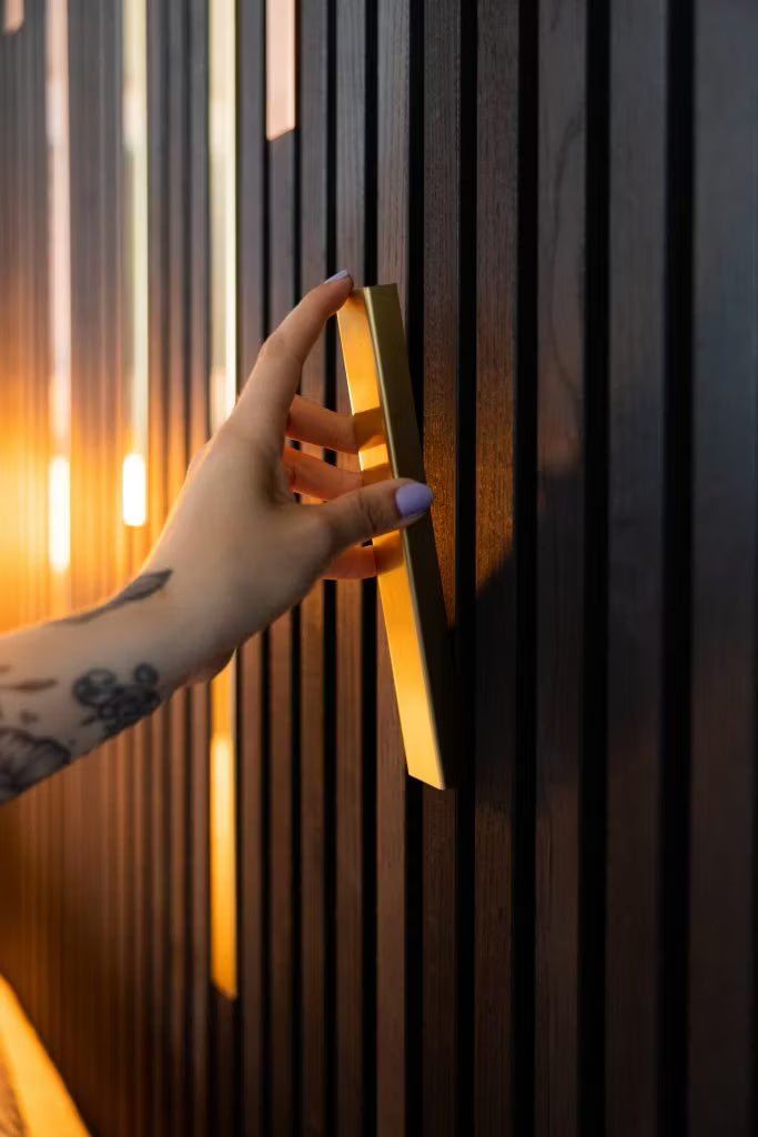 Eine tätowierte Hand mit hellvioletten Nägeln hält einen goldenen Griff an einer glatten, dunklen Akupanel-Holzpaneelwand, die von einer warmen Beleuchtung hervorgehoben wird.