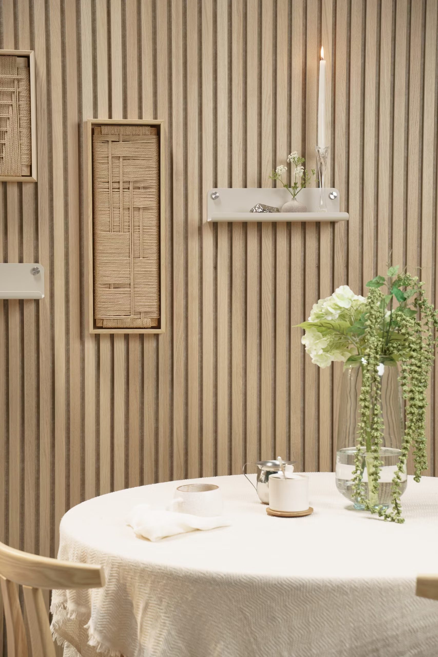 Ein runder Tisch mit einer hellen Tischdecke, frischen Blumen in einer Vase und einer Kerze steht vor einer Akupanel-Akustikholzlattenwand im nordischen Stil mit minimalen Regalen.