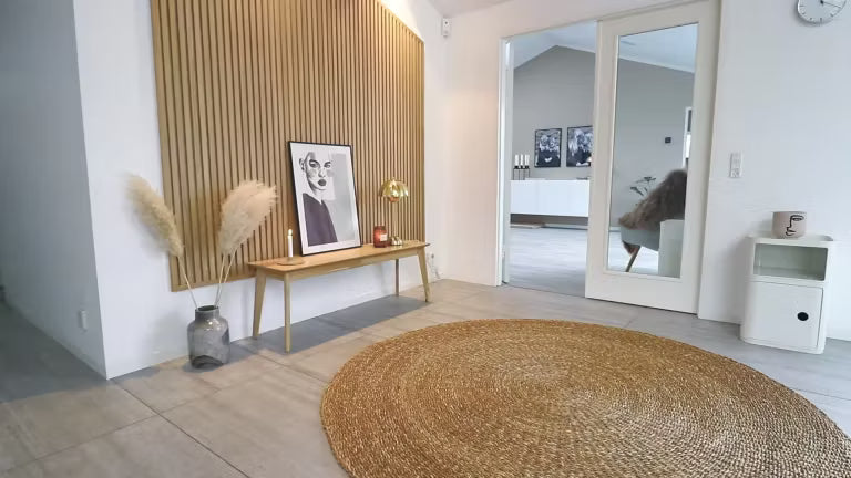 Minimalistischer nordischer Wohnraum mit einem runden Juteteppich, einer Holzbank mit Pampasgras, gerahmter Kunst und einer Akupanel-Akustikholzwand mit Lattenrost.