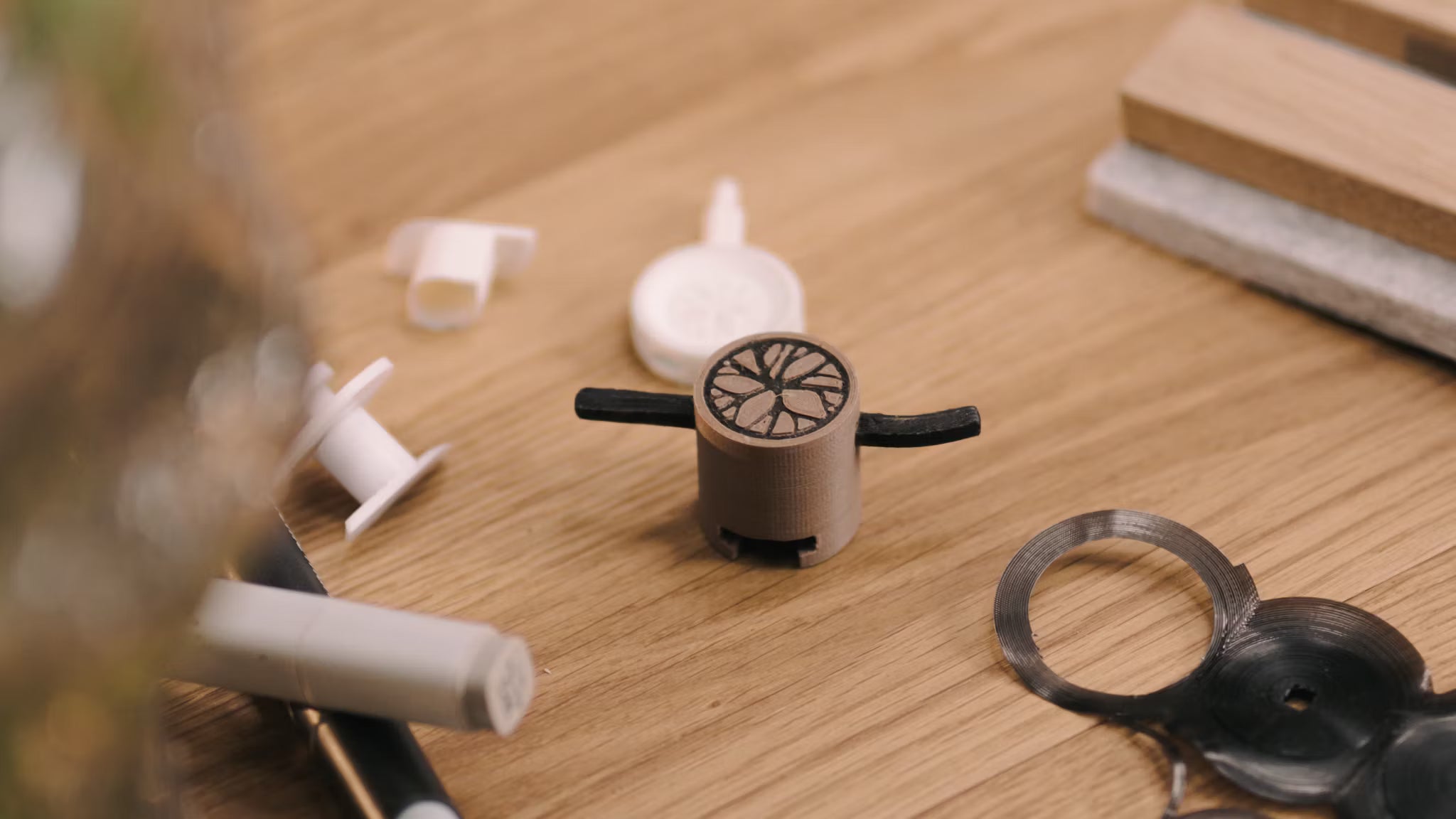 Ein kleiner 3D-gedruckter Prototyp mit schwarzen Armen liegt auf einem Holztisch neben verschiedenen Kunststoffkomponenten und verweist auf die Entwicklung von Zubehör für Akustikplatten.