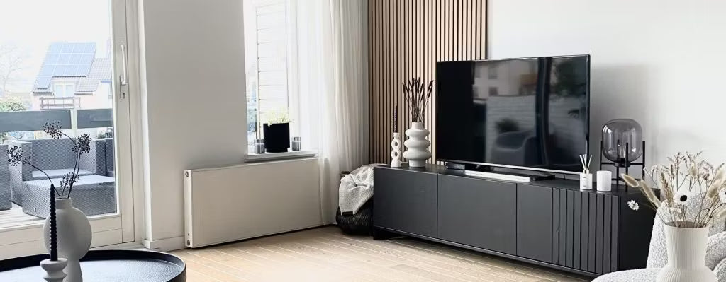 Modernes Wohnzimmer mit schwarzem TV-Ständer, Flachbildfernseher, dekorativen Vasen und großen Fenstern mit Tageslicht und Blick auf den Innenhof; Akupanel an einer Wand.