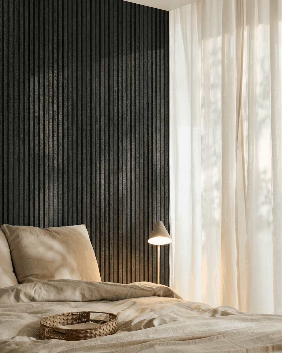 Gemütliches Schlafzimmer mit beige Bettwäsche, einem Tablett aus Rattan, weißer Lampe, transparenten Vorhängen und einem WoodUpp Akupanel Akustikholzpaneel in Charcoal, das an der Wand montiert ist.