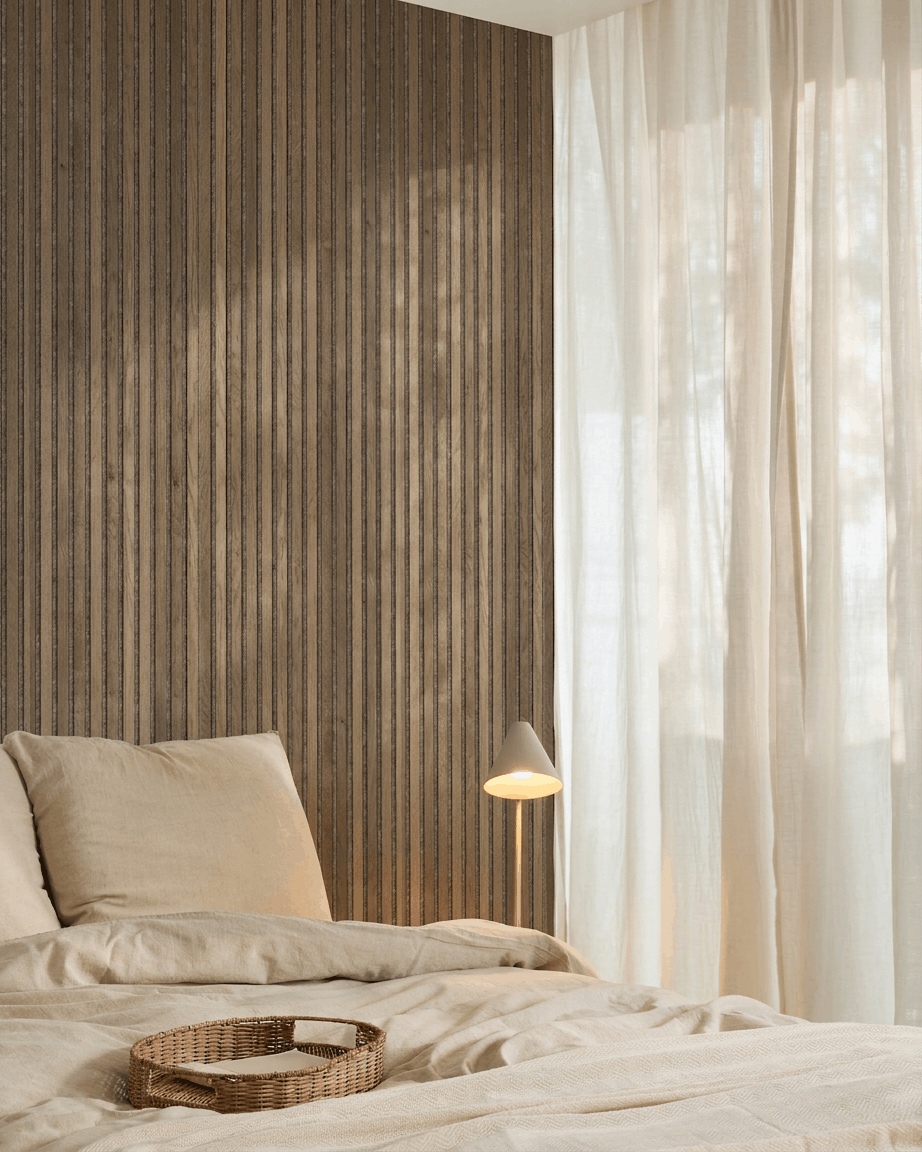 Minimalistisches Schlafzimmer mit beige Bettwäsche, einem Tablett aus Rattan, einem WoodUpp Akupanel Rustic Grey Oak Akustik-Lamellenwandpaneel, einer weißen Lampe und transparenten Vorhängen.