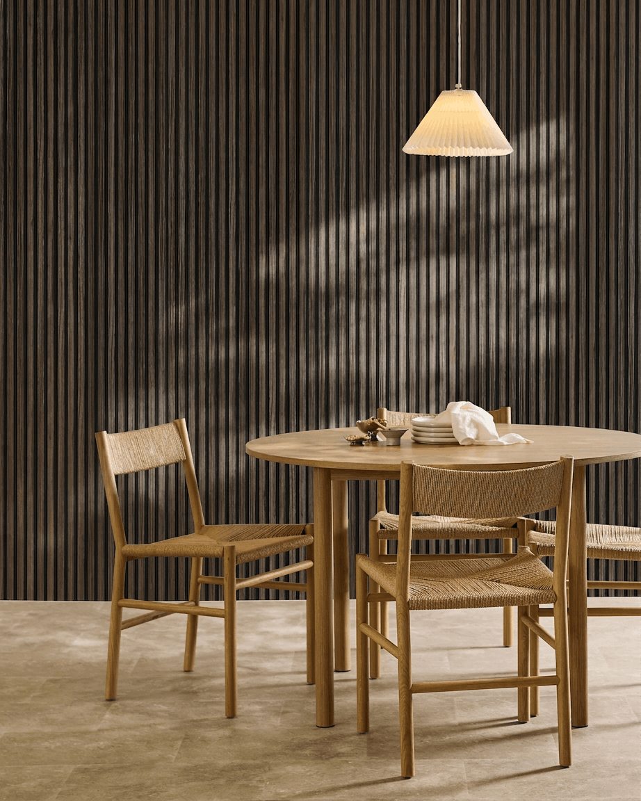 Ein runder Tisch mit vier geflochtenen Stühlen steht neben einem WoodUpp Akupanel Black Oak Akustikpaneel unter einer plissierten Pendelleuchte.